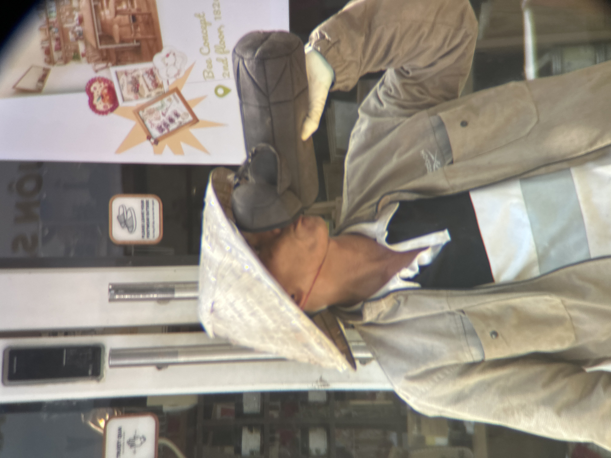 A person wearing a conical hat and a light-colored jacket is holding a dark, cylindrical object to their mouth, appearing to drink from it. The person is standing in front of a building with glass doors and windows. A sign in the background reads "Bee Concept, 2nd floor, 1826". The overall scene suggests an outdoor setting in a city, likely in Đà Nẵng, Vietnam, given the conical hat and the likely context of the sign.