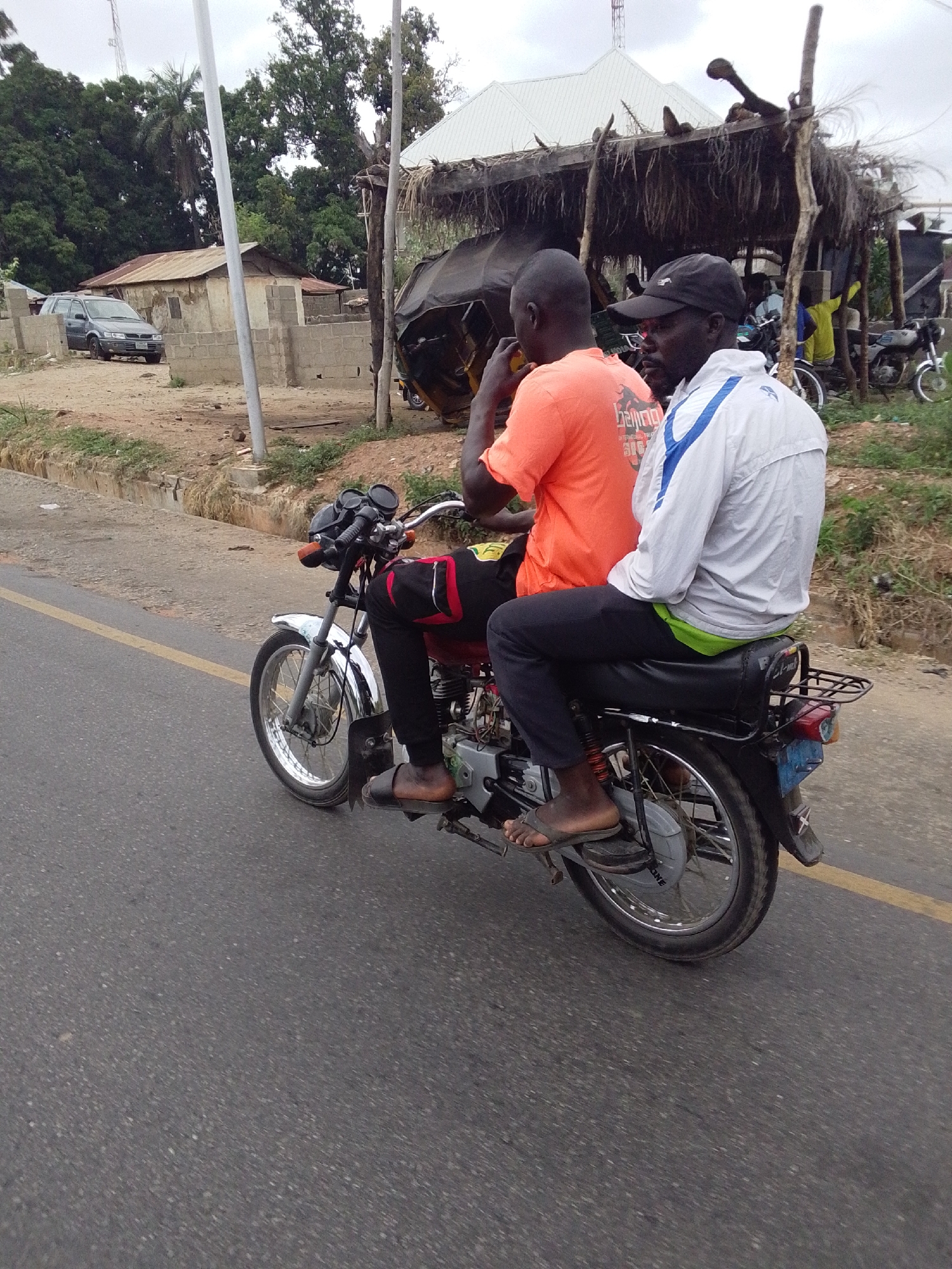 Two men ride a motorcycle on a paved road. The driver is wearing an orange t-shirt, black pants with red and white accents, and sandals. The passenger is wearing a white jacket with blue accents, dark pants, and a black baseball cap. The motorcycle is red and silver with black accents. A yellow line is visible on the right side of the road. In the background, there is a small building with a corrugated metal roof, a thatched-roof structure, and trees. A car is parked in front of the building. The location is likely in Lokoja, Nigeria.