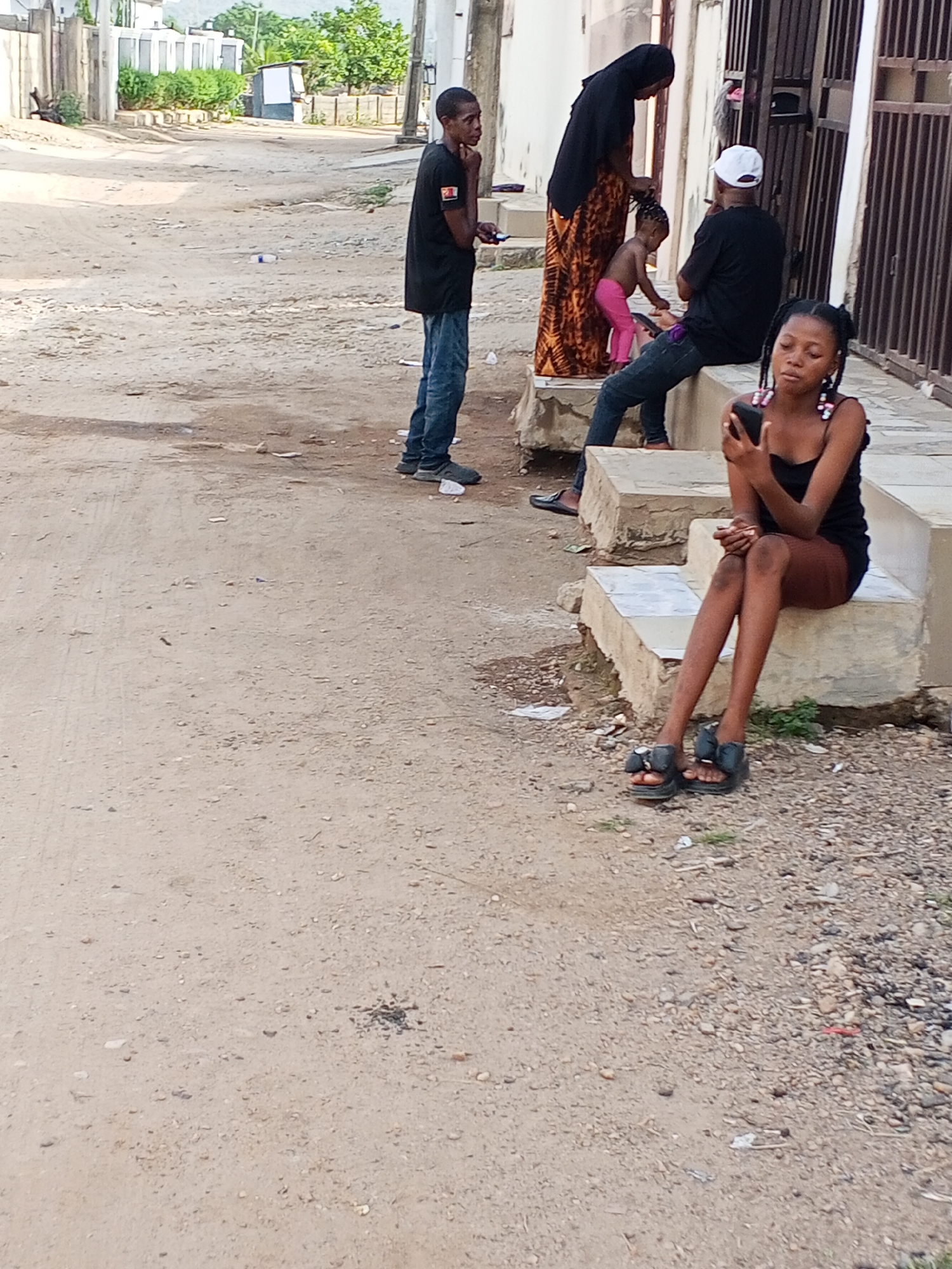 The image shows a street scene in Otokiti, Nigeria. In the foreground, a young woman with braided hair sits on concrete steps, looking at her mobile phone. She wears a black top, a dark skirt, and black sandals.
In the background, several other people are present. A young man stands on the street, looking towards a group seated on another set of concrete steps. This group includes a woman wearing a black hijab and an orange patterned dress, a toddler in pink clothing, and a man wearing a white cap and black shirt. All are outside of a building.
The street is unpaved and covered in a mix of dirt and gravel. Trash litters the steps and the ground. Buildings and foliage are visible in the background. The sky appears overcast.