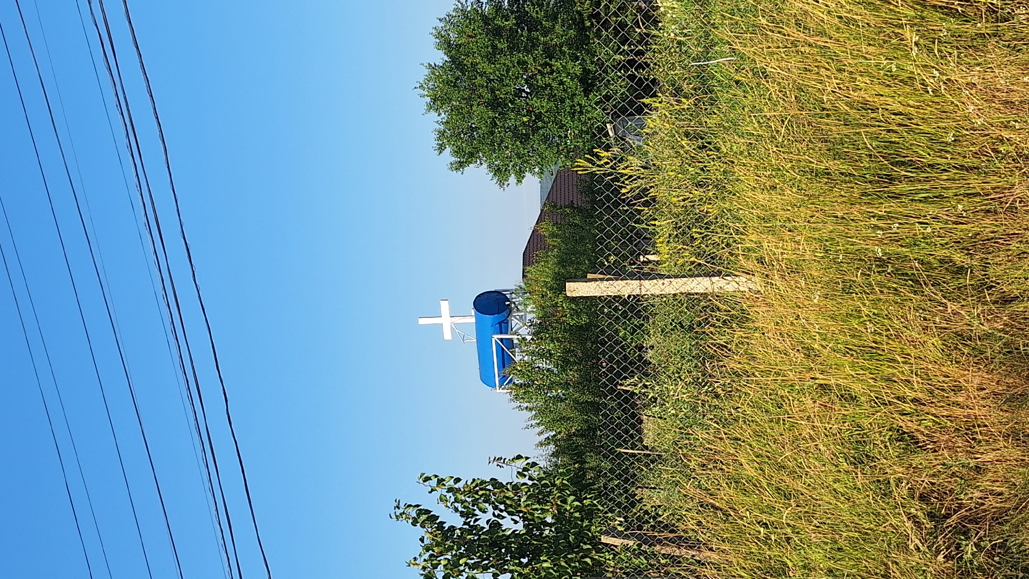The image shows a rural scene with a water tower topped by a white cross, set against a clear blue sky. The water tower is a large, cylindrical blue tank supported by a metal structure. The cross is white and prominently placed on top of the tank. In the foreground, a chain-link fence runs horizontally across the frame, separating the viewer from a grassy field with tall, dry grass and some scattered green vegetation. Behind the fence and the water tower, there are trees and what appears to be a building with a dark roof. Overhead, several power lines are visible against the sky. The scene is likely located in a village or rural area, given the presence of the water tower, the fence, and the vegetation. Sărata-Galbenă is a village in Moldova, which aligns with the appearance of the scene. There are no people visible in the image.