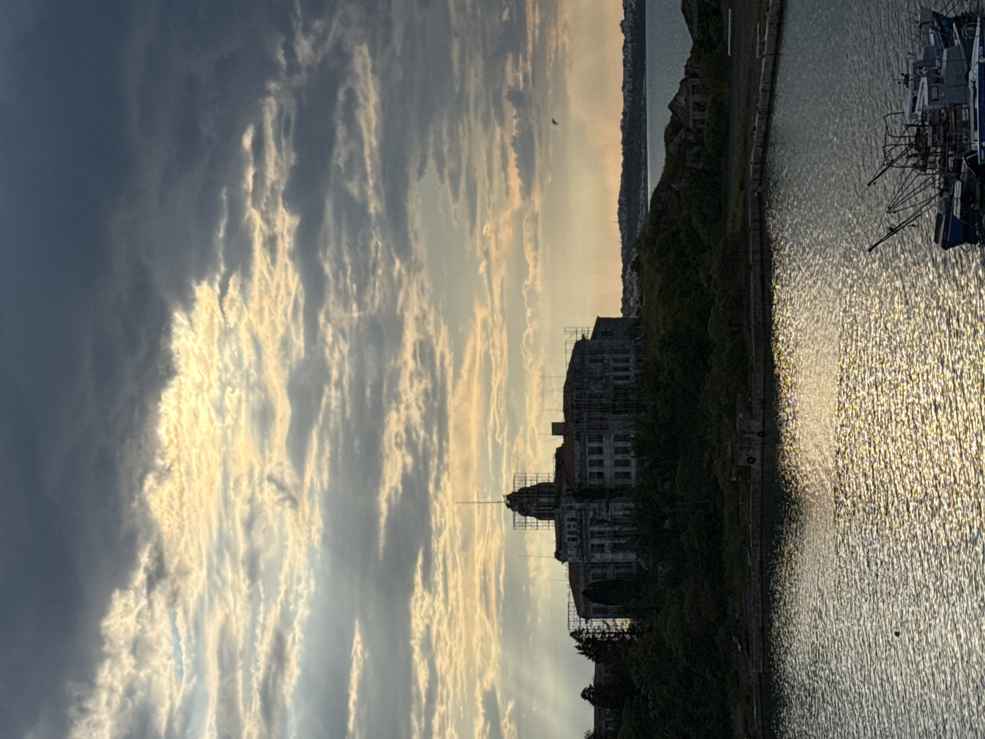 The image captures a waterfront scene in Sozopol, Bulgaria, likely taken at dawn or dusk given the golden light filtering through the clouds. The sky is predominantly cloudy, with streaks of light breaking through. In the foreground, there is a body of water, possibly the Black Sea, with shimmering reflections of light on its surface. A harbor wall and a line of dense trees separate the water from a dilapidated multi-story building under scaffolding. The building's structure is partially visible, suggesting that renovations are underway. In the far right of the frame, partially cropped, are some boats.