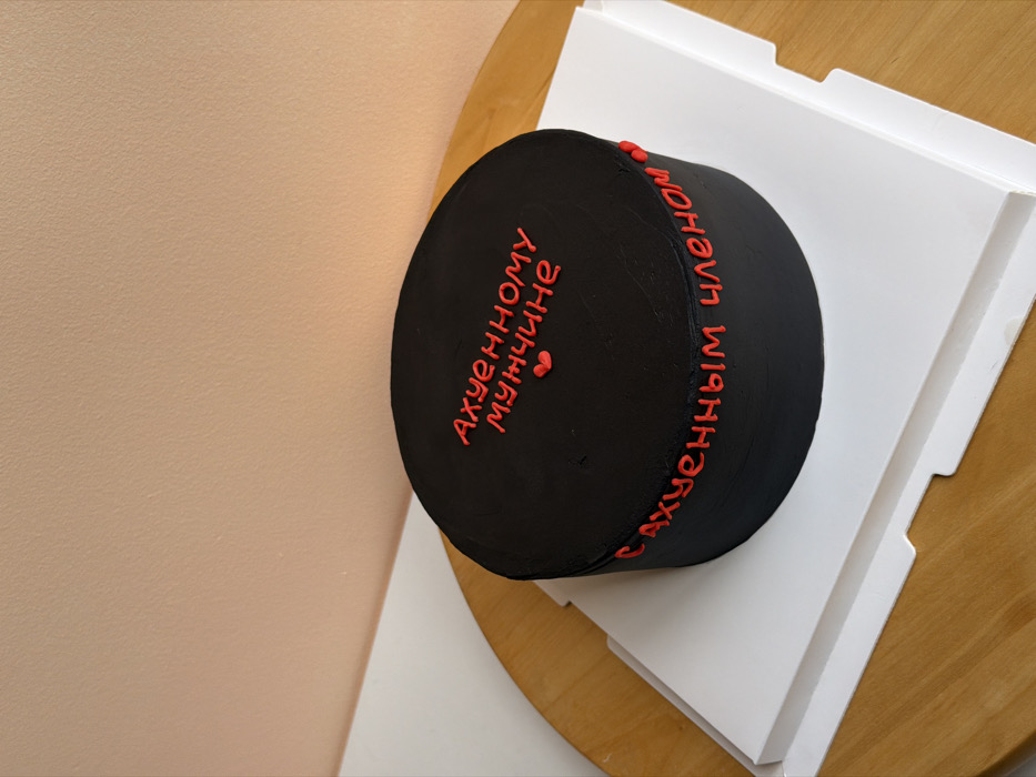 A cylindrical cake, frosted entirely in black, is centered on a white square cardboard cake base. Red icing text adorns the cake: "Ахуенному мужчине" (To an awesome man) is piped across the top, accompanied by two small red hearts. The phrase "С ахуенным членом" (with an awesome member/penis) is piped around the vertical side of the cake. This assembly rests on a light-toned wooden surface, likely a table, which occupies the lower portion of the frame. The background consists of a plain, smooth, light peach or beige colored wall, occupying the upper two-thirds of the image. No individuals, explicit actions, or interactions are visible. The scene is indoors. No visual elements within the image provide evidence to identify the location as Tel-Aviv, Israel.