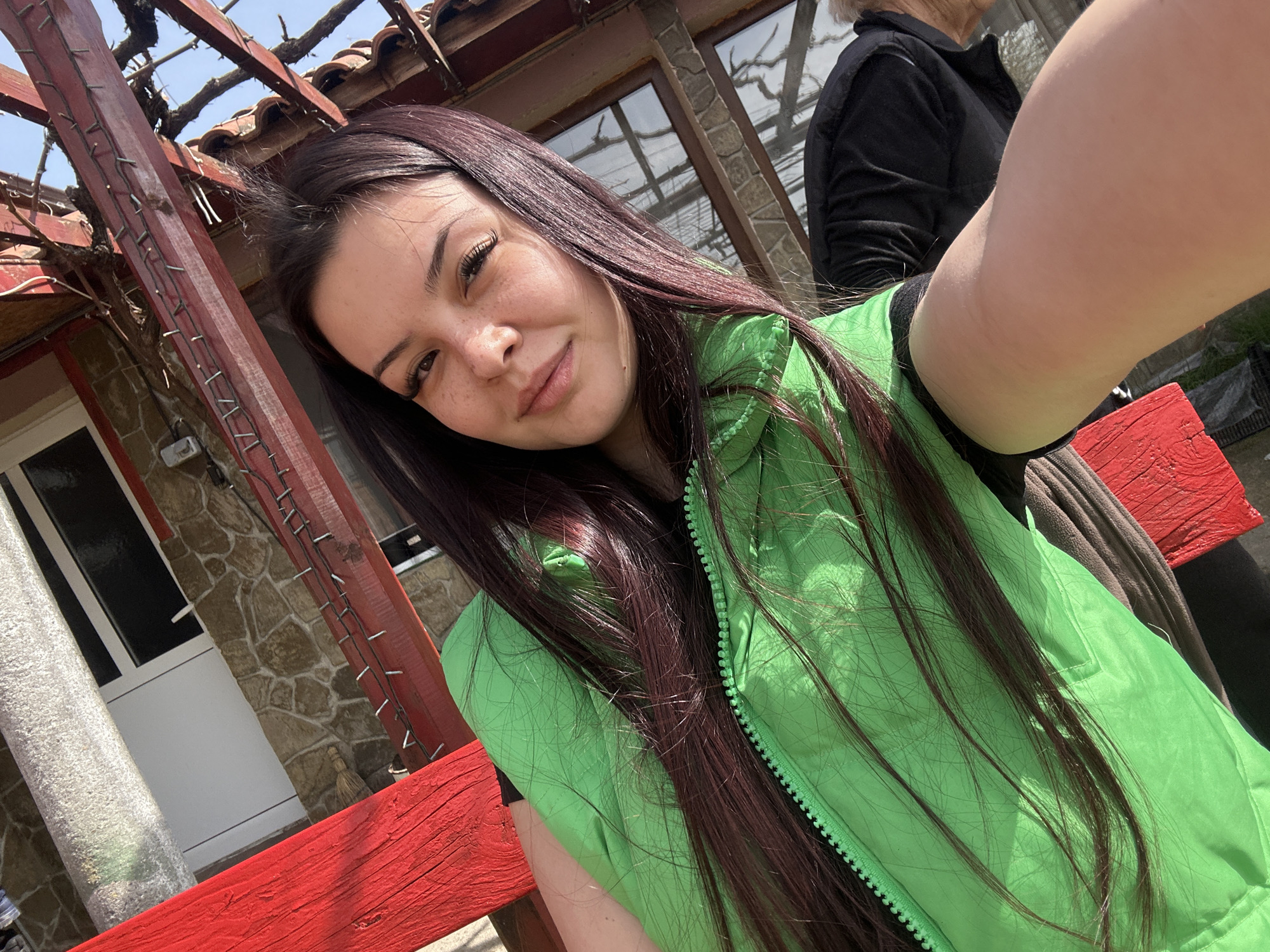 The image is a selfie taken by a young woman with long, dark auburn hair. She is wearing a bright green vest. Behind her, partially visible, is another person wearing a black jacket. The background features a building with stone siding and white trim around a door or window. A wooden pergola structure with red-painted beams is also visible. The location appears to be outdoors in Chubra, Bulgaria.
