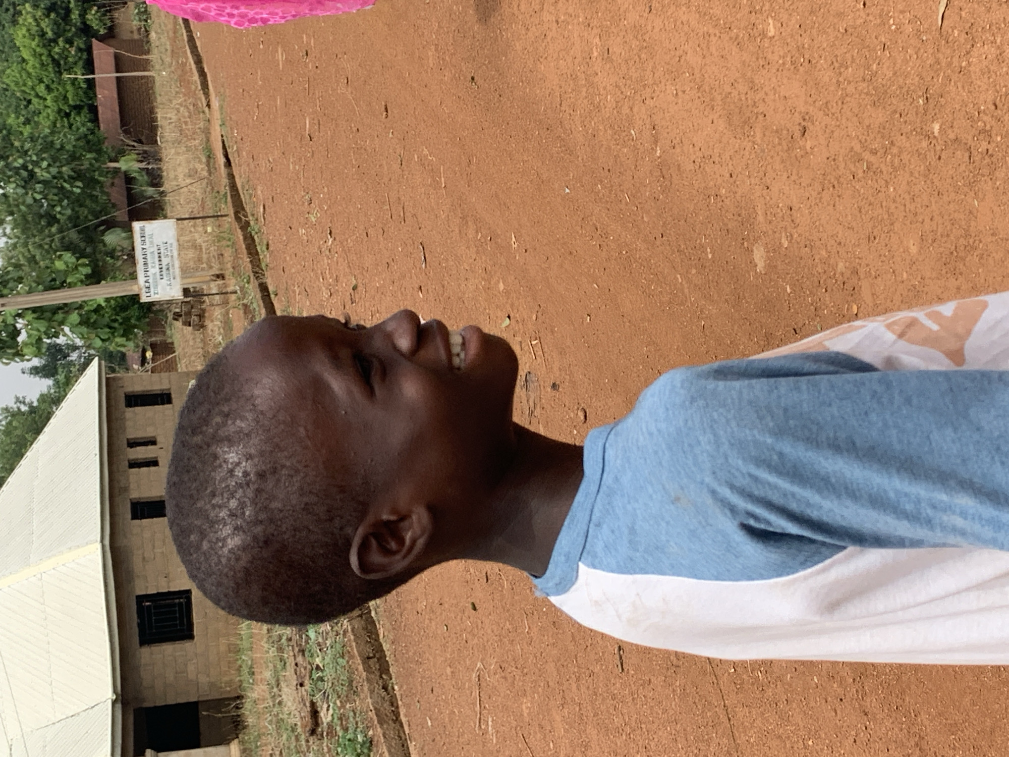 The image shows a person in Agban, Nigeria, standing outside near a building. The person is wearing a light blue and white shirt. To the left of the person is a building with a metal roof and dark-framed windows. In the background, a sign for "L.G.E.A. Primary School Zuntu" is visible. The ground is reddish-brown.