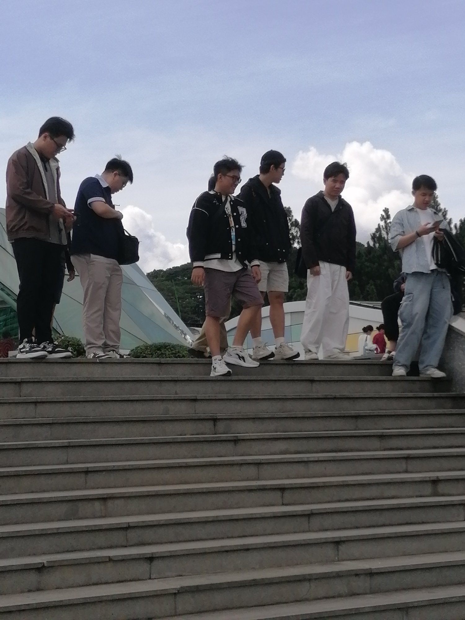 A group of seven people are standing on a set of outdoor stairs. The people are of varying ages and are dressed casually. The stairs appear to be made of concrete and are surrounded by greenery. The background shows a cloudy sky and some trees. One of the people is holding a phone and looking at it. Another person is looking down at the stairs. The scene appears to be a public space, possibly a park or a tourist attraction. The architectural style of the building in the background, with its triangular glass panels, suggests it could be the Da Lat Railway Station, a historic landmark in Da Lat, Vietnam. The station is known for its unique architecture, which incorporates elements of both French colonial and indigenous Vietnamese styles. The overall atmosphere of the image is casual and relaxed, with people enjoying their time outdoors.