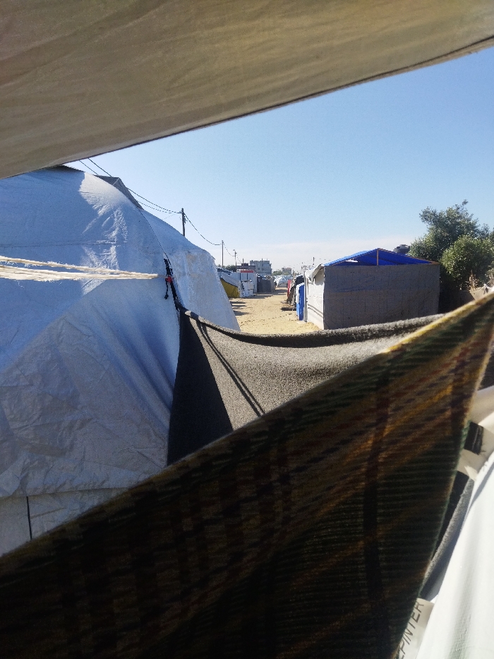 The image depicts an outdoor encampment scene in Khan Yunis, Palestinian Territory, viewed from underneath a light-colored fabric canopy that occupies the top portion of the frame. In the immediate foreground, a dark, patterned blanket hangs diagonally across the lower right. To its left, a dark grey fabric is also visible. The midground on the left features a large, light-colored tent, likely constructed from tarp material, with visible ropes and dark fastening clips. A sandy or dirt pathway extends into the background, flanked by various temporary structures. On the right side of the pathway, a rectangular structure with a blue tarp roof and rough-textured walls is visible, accompanied by green foliage. Further down the pathway, an assortment of other tents and structures, including white, yellow, and blue elements, are present. Overhead power lines with poles run along the pathway. In the far distance, several permanent-looking buildings and a line of vehicles are discernible. The sky is clear and blue. No individuals or specific actions are evident within the frame.