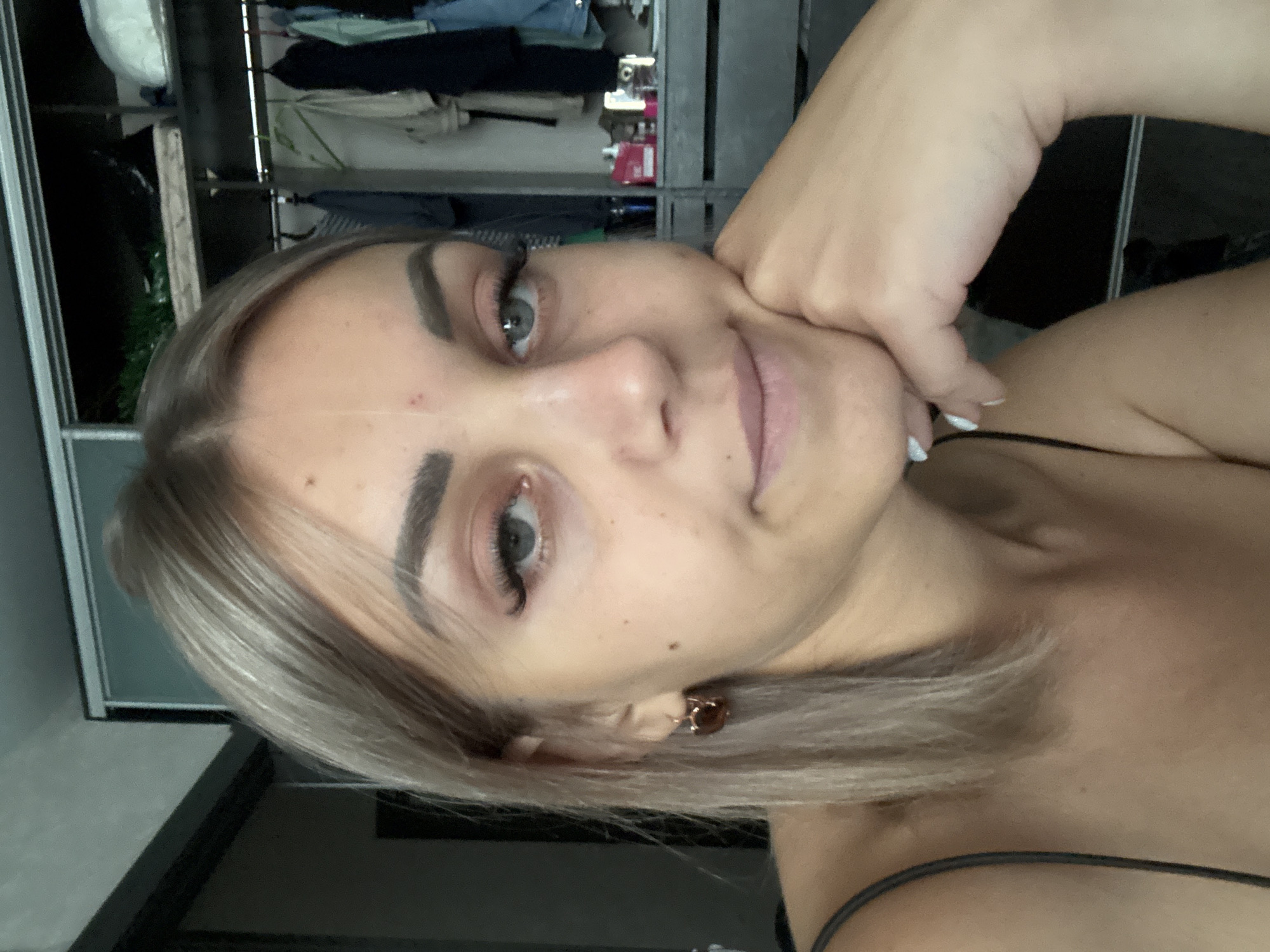A young Caucasian woman with blonde hair and light blue eyes is shown in a close-up selfie. She is wearing a dark tank top and a small, brown earring. Her right hand is propped under her chin, and her left hand is resting on her cheek. The background appears to be a closet or wardrobe area with clothing on hangers. The image was taken indoors, possibly in a bedroom or dressing area. No specific details about the city of Yuzhno-Sakhalinsk, Russia, can be inferred from the visual information provided.