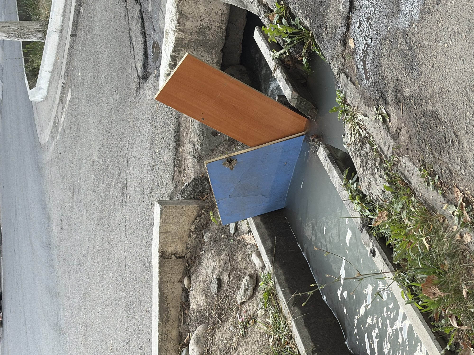 Two pieces of furniture, a blue door and a wooden plank, are partially submerged in a drainage ditch filled with murky water. The blue door is propped up at an angle, while the wooden plank leans against it, also partially submerged. The drainage ditch is concrete-lined and surrounded by rough terrain with scattered rocks and sparse vegetation. An asphalt road with visible cracks and patches is visible in the background, along with a concrete curb. The scene appears to be an outdoor urban environment.