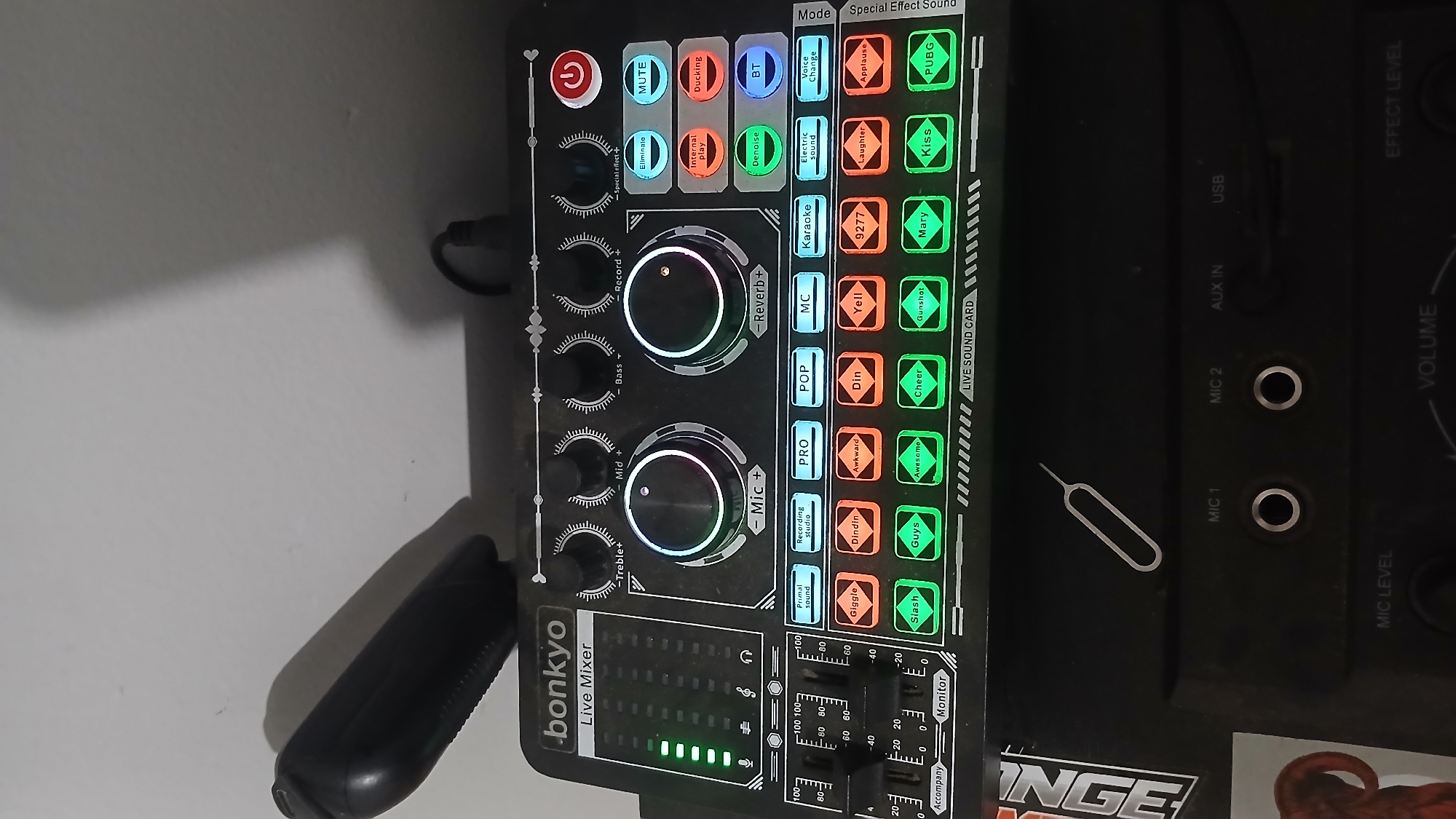 The image shows a close-up, slightly angled, eye-level view of a Bonkyo Live Mixer device situated on top of another black electronic device. The mixer is black with various knobs, buttons, and illuminated LED lights. The "Bonkyo Live Mixer" branding is visible on the top left. The device has a power button on the top right. Various buttons with labels like "Primal sound," "Recording studio," "PRO," "POP," "MC," "Karaoke," "Electric sound," "Voice Change," "Giggle," "Dindin," "Awkward," "Din," "Yell," "9277," "Laughter," "Applause," "Slash," "Guys," "Awesome," "Cheer," "Gunshot," "Mary," "Kiss," and "PUBG" are arranged in rows. There are also controls for "Treble," "Mid," "Bass," "Reverb," and "Mic." A set of green LED level indicators are on the left side. Below the mixer, a SIM card ejector tool is lying on the black surface of the other electronic device, which has ports labeled "MIC 1," "MIC 2," "AUX IN," and "USB," along with "MIC LEVEL," "VOLUME," and "EFFECT LEVEL" controls. The background is a plain, light-colored wall with a shadow cast by the mixer and a microphone extending from the top left. The scene appears to be indoors, possibly in a home or studio setting. The location in Sampang, Indonesia, is not visually identifiable from the image content.
