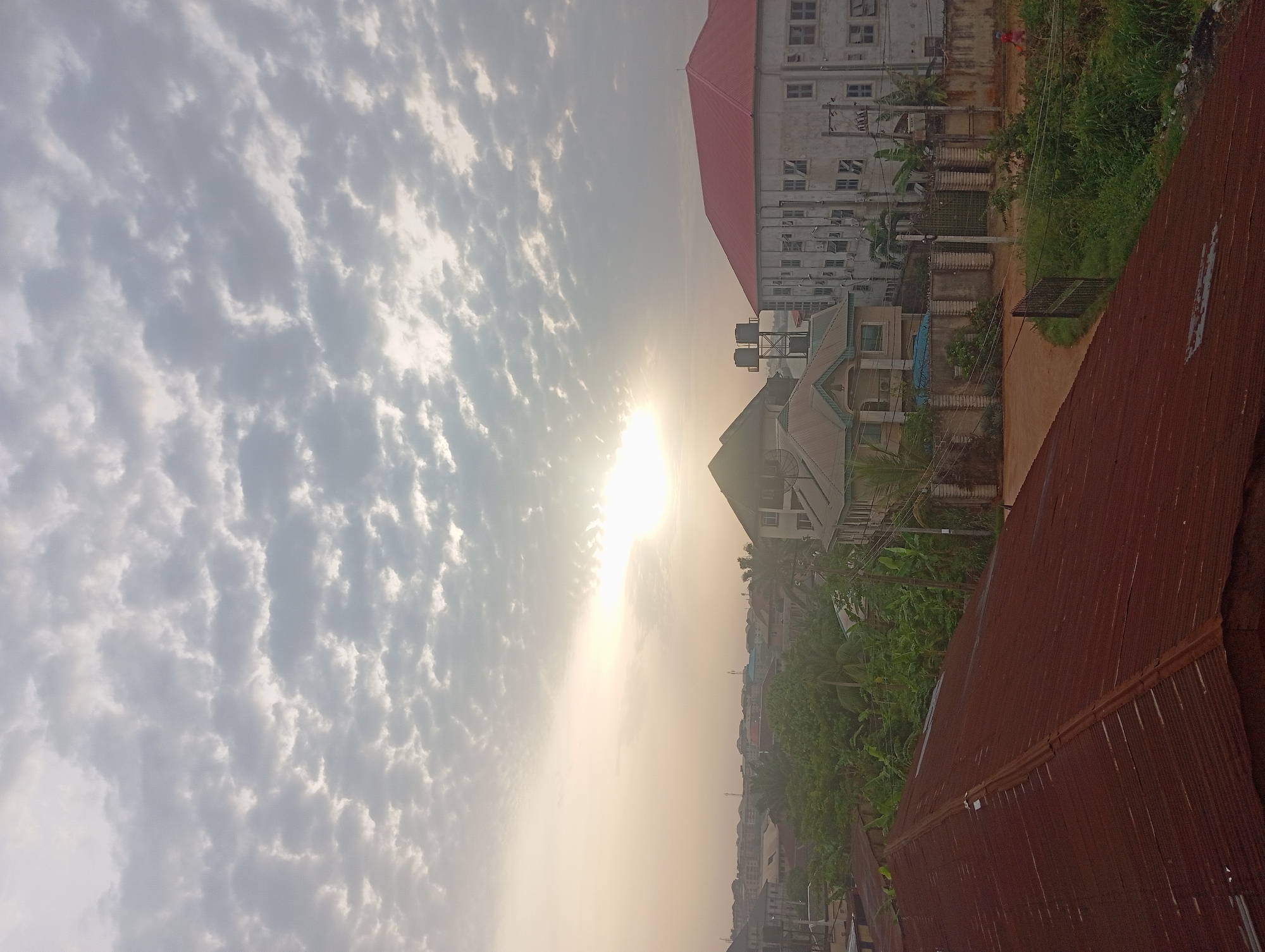 The image captures a daytime urban scene in Nkpor, Nigeria, featuring a hazy, sunlit sky with scattered cloud formations. The foreground includes a corrugated metal rooftop. In the midground, there are several buildings of varying sizes and architectural styles, some painted white with red roofs, alongside vegetation, power lines, and water tanks. The environment exhibits signs of urbanization with residential and commercial structures. The scene is viewed from an elevated perspective.