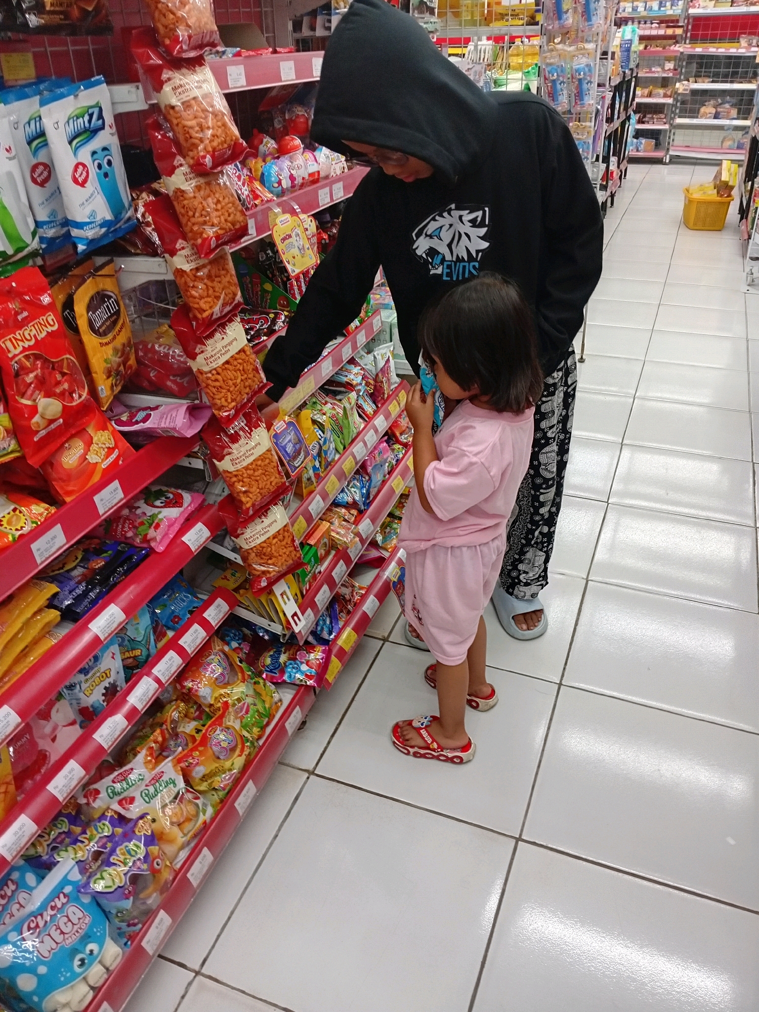 A person wearing a black hoodie and patterned pants stands in an aisle of a brightly lit convenience store, with a young child in a pink t-shirt and shorts standing beside them. The child is looking at a package of snacks on a red shelf. The shelves are stocked with various packaged goods, including candy, chips, and drinks. The floor is tiled with white square tiles. A yellow basket is visible on the right side of the aisle. The store appears to be located in Seren, Indonesia, indicated by the presence of products with Indonesian labeling.