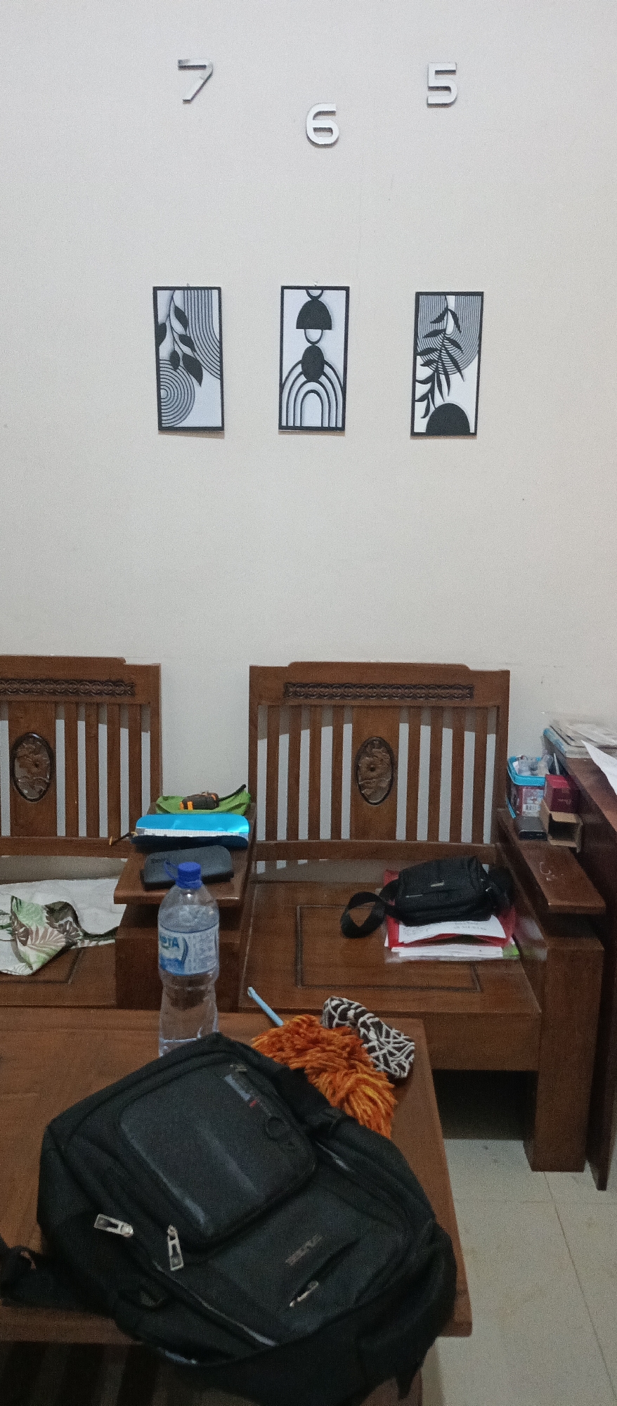 In a room with a neutral-colored wall, three framed black and white abstract prints are displayed vertically. Below the prints, two wooden chairs, possibly part of a sofa, are arranged side-by-side. The chairs feature vertical slatted backs with decorative circular inlays. On the seat of the left chair, a green pouch, a blue pencil case, and a grey pouch are placed. A bottle of "AQUA" brand mineral water stands on a wooden surface between the two chairs. To the right of the water bottle, an orange duster with a patterned fabric is visible. In the foreground, a black backpack with multiple zippered compartments rests on a wooden surface. The scene appears to be an interior space, potentially a living room or a common area, in Arjasa, Indonesia.