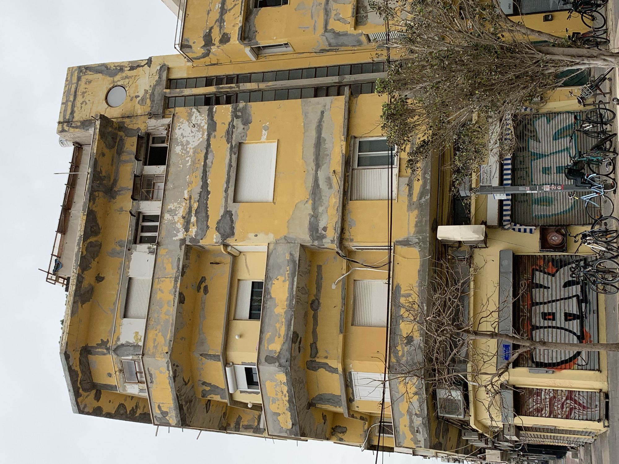The image shows an old, yellow building in Tel-Aviv, Israel. The paint is peeling off, revealing a grey underlayer. The building has several windows, some with closed blinds. A tree is in front of the building, partially obscuring the view. A closed storefront with graffiti and bicycles parked in front of it are next to the building. The sky is overcast.