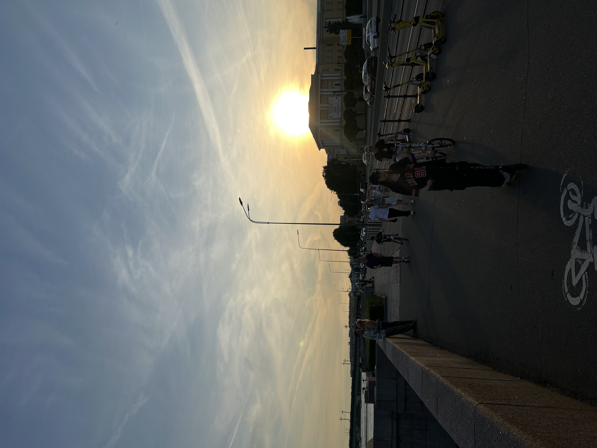 The media file depicts a sunset scene in Saint Petersburg, Russia. A bright sun, partially obscured by wispy clouds, hangs in the sky, casting a warm, golden glow over the cityscape. The sky is a mix of blue and yellow hues, with long, streaky clouds suggesting a gentle breeze.
In the foreground, a paved walkway runs alongside a body of water, likely a canal or river. People are engaged in various activities: some are walking, while others are riding electric scooters and bicycles. A group of yellow electric scooters are parked in a row, suggesting a rental service. A woman on a scooter in the foreground wears a black shirt with "16 NYT SUT" visible in red lettering. Another woman in the background appears to be resting on a stone ledge overlooking the water.
The scene includes a building with a classical architectural style, likely in the background. Streetlights line the walkway, indicating an urban setting. The overall mood is relaxed and pleasant, with the warm lighting of the sunset contributing to a serene atmosphere. The image captures a typical evening in the city, with residents enjoying outdoor activities as the day winds down.