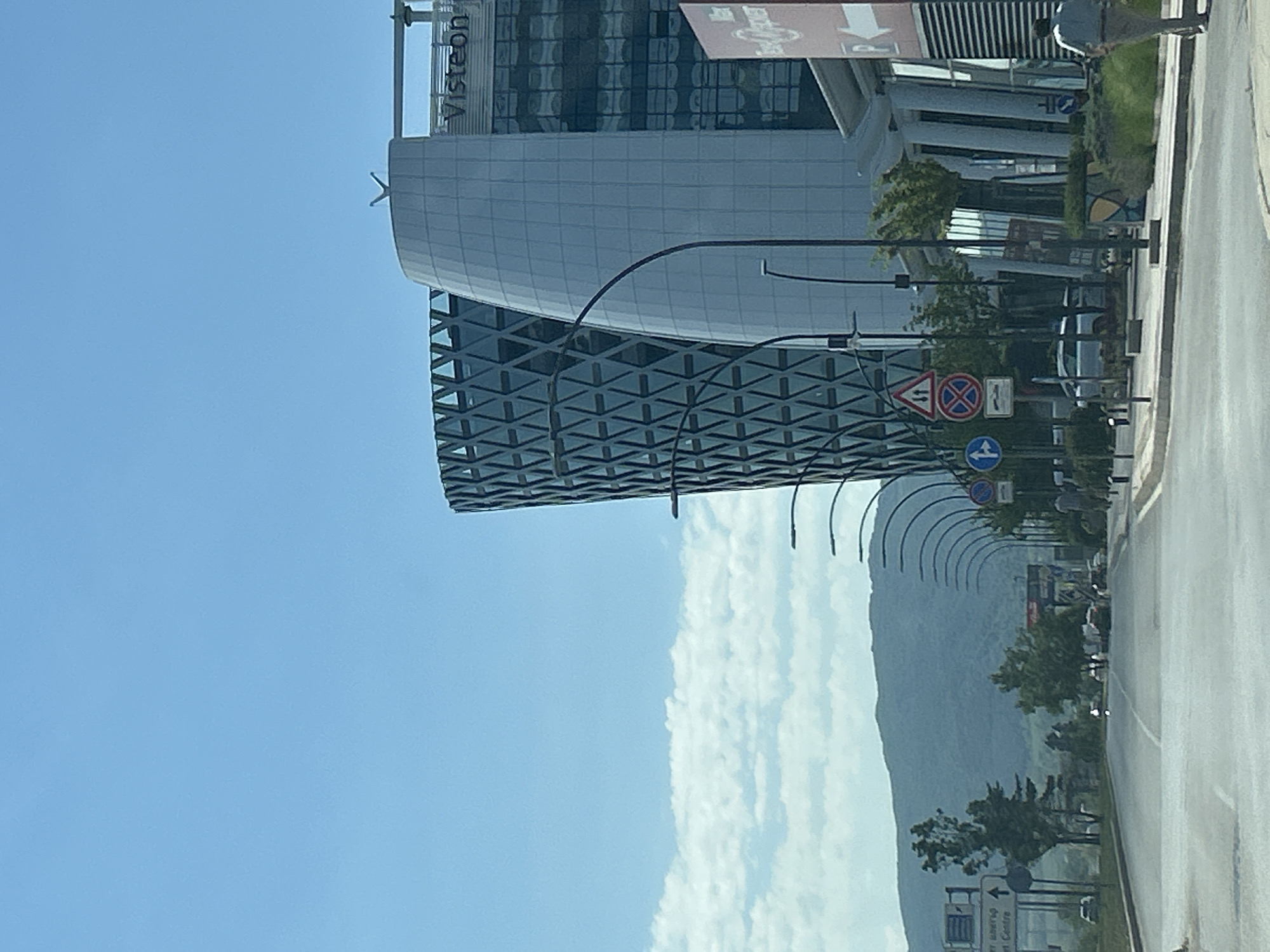 The image shows a street scene in Sofia, Bulgaria, featuring modern architecture and urban infrastructure under a clear blue sky. In the background, a large, contemporary building with a distinctive curved facade dominates the skyline, bearing the "Visteon" logo. To its left is another building with a complex geometric pattern on its exterior. The street is lined with lampposts and various traffic signs, including directional signs and regulatory symbols like a double-arrow sign and a circular sign with a red cross. Trees and greenery are visible along the roadside, with hills or mountains forming a natural backdrop in the distance. Several cars are present on the road, and a few people are seen walking on the sidewalk. The overall scene depicts a well-developed urban area with a blend of commercial and natural elements.