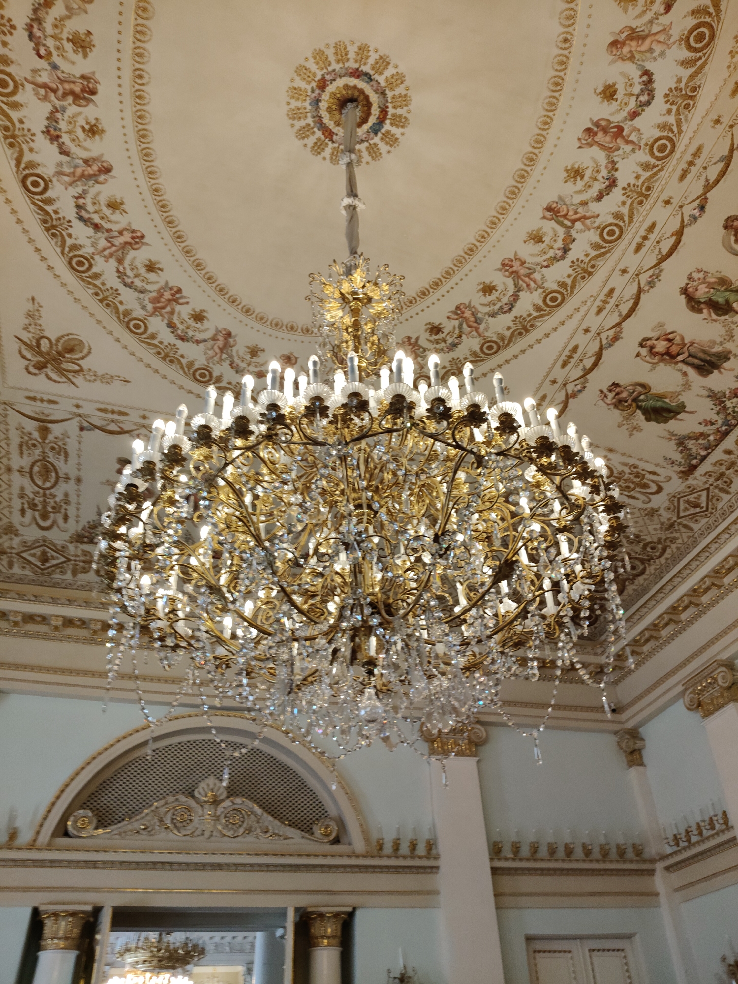 A crystal and gold chandelier hangs from a richly decorated ceiling in a grand room. The ceiling is adorned with painted cherubs and ornate gold detailing. Below the chandelier, a semicircular architectural feature with intricate gold ornamentation is visible, above what appears to be a doorway or archway. The walls are painted a pale color, with further gold decorative elements and a series of sconces holding candles. The room's opulence suggests it could be a ballroom, reception hall, or a similar space within a historical palace or museum. Based on the style and common depictions of opulent interiors in Saint Petersburg, this is likely the Catherine Palace in Pushkin, near Saint Petersburg, Russia, possibly within the Amber Room or a similar gilded chamber.