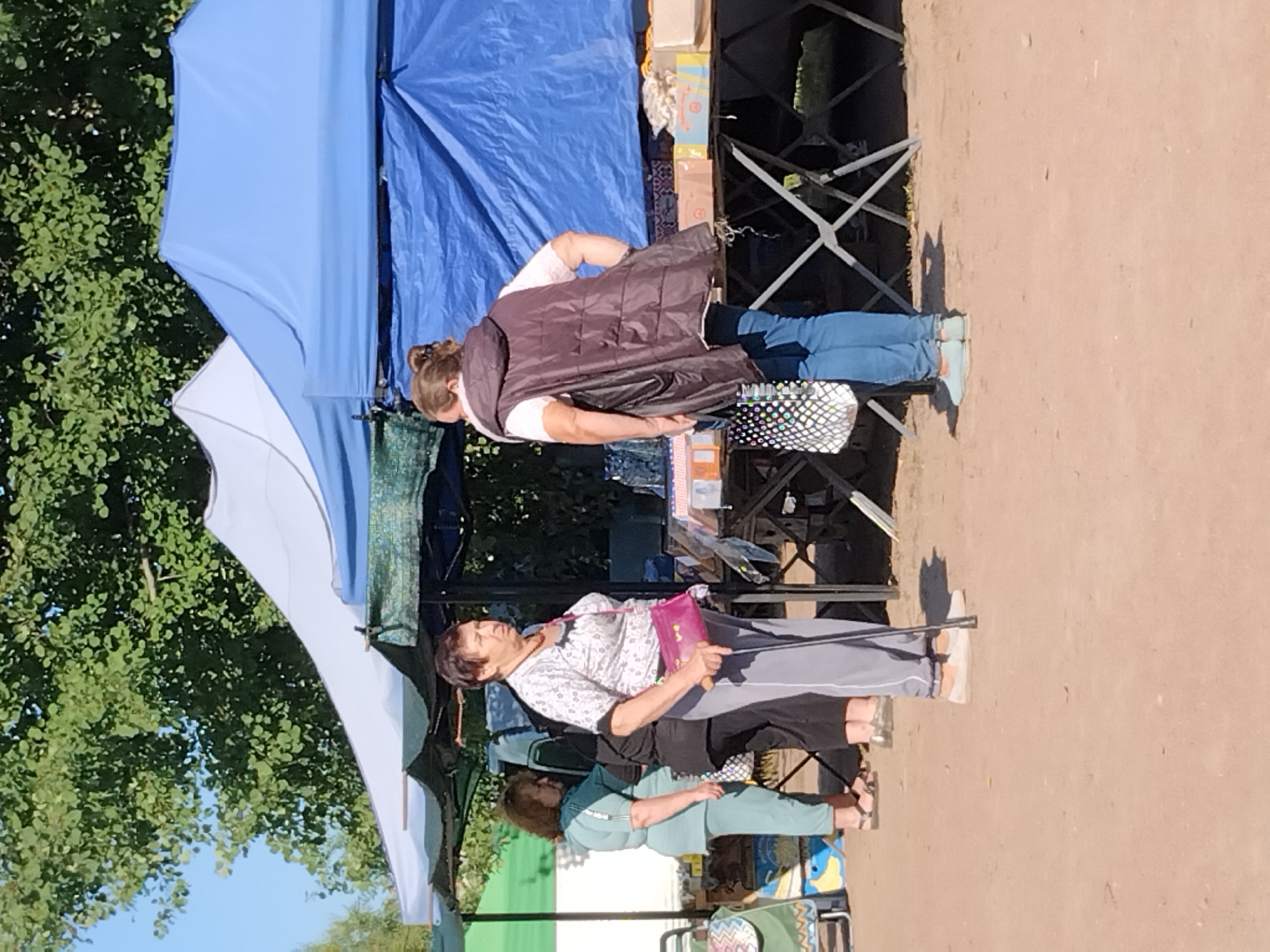 A scene at an outdoor market in Irshansk, Ukraine, features several individuals under blue and white tents. In the foreground, a woman with short dark hair, wearing a patterned shirt and gray pants, holds a cane and a magenta purse. She stands facing a woman with brown hair tied back, who is wearing a white shirt, a dark vest, and blue jeans, and is holding a patterned bag. To the left, a woman in a teal tracksuit stands near a shopping cart. In the background, stalls display various items, and lush green trees are visible overhead. The ground is covered with a light brown, sandy material. The time of day appears to be sunny and warm.