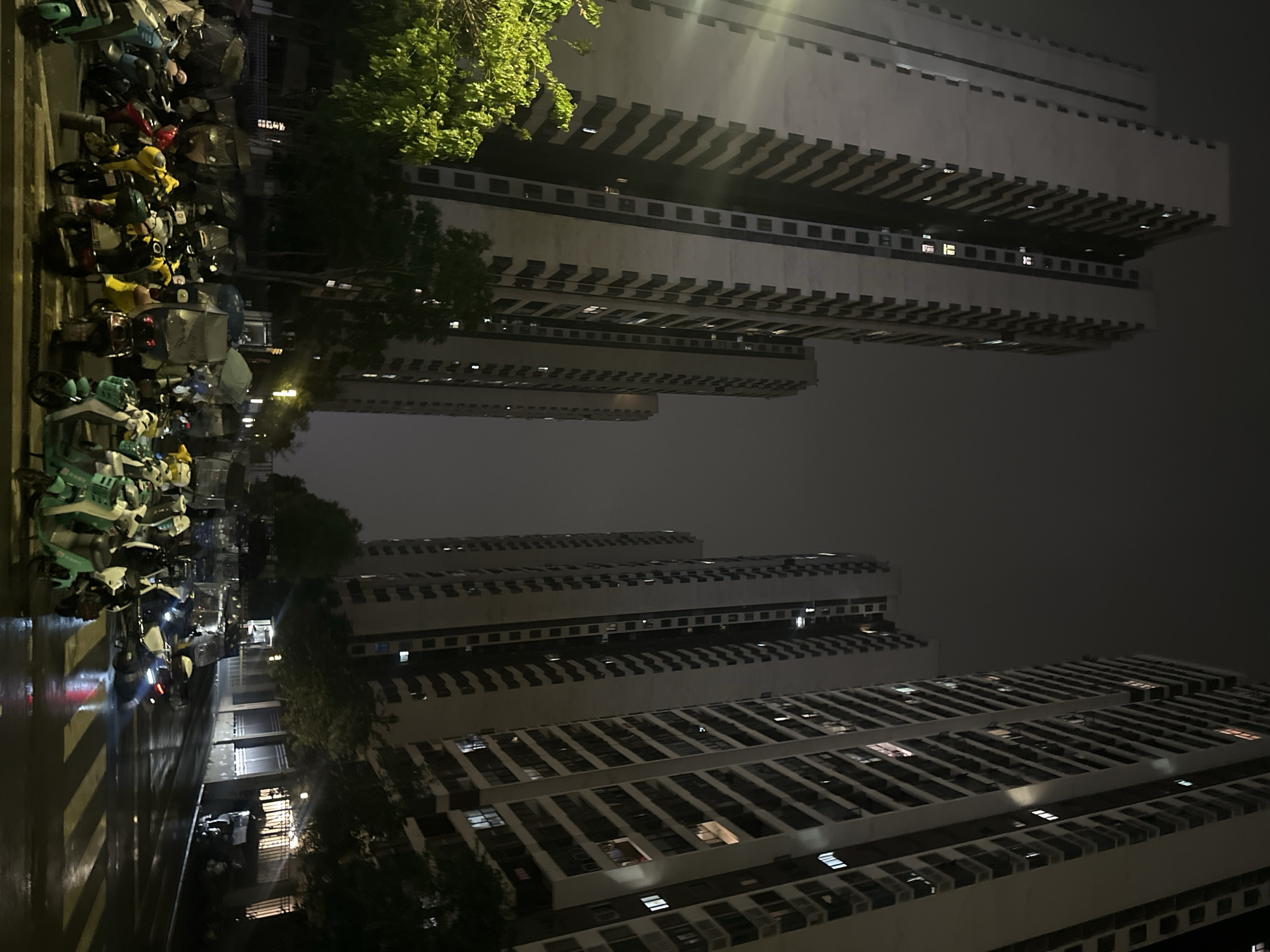 The image depicts a nighttime scene in the Chongqing High-tech Zone, China, showing apartment buildings and a street with parked motorcycles. The apartment buildings are tall with many windows, some illuminated. The street is wet, reflecting the city lights, and lined with parked motorcycles and bicycles. The sky is overcast and dark.