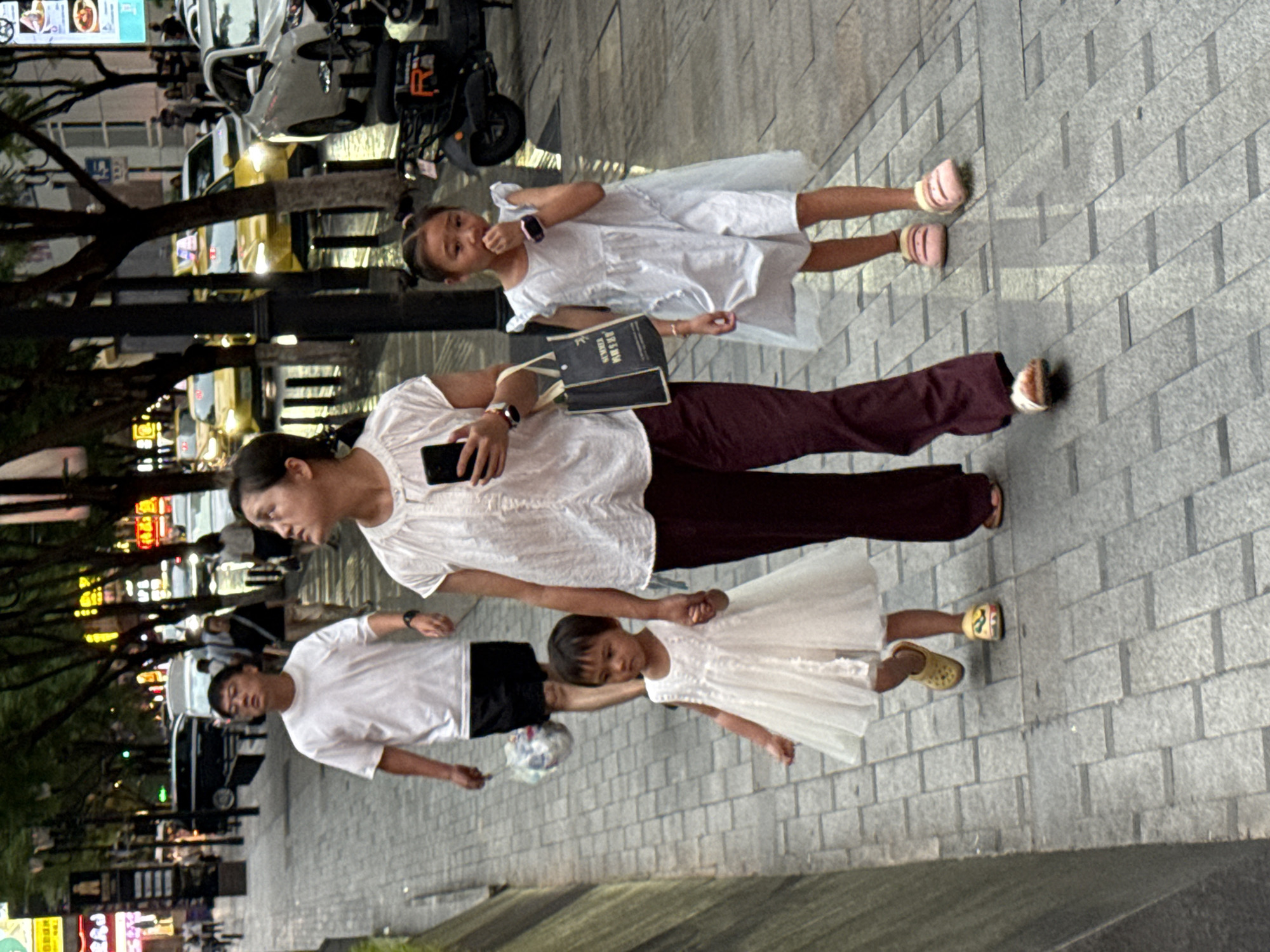A family walks down a brick-paved sidewalk in Yuzhong District, China. A man in a white t-shirt and black shorts walks ahead, carrying a plastic bag. A woman in a white top and burgundy pants walks behind him, holding a smartphone in her right hand and a dark tote bag in her left. Two young girls walk beside the woman. The younger girl, in a white dress, walks to the woman's left, holding her hand. The older girl, in a light blue dress, walks to the woman's right. The sidewalk is lined with trees. In the background, cars are visible on a street, including yellow taxis and white vehicles. Buildings and streetlights are also present.
