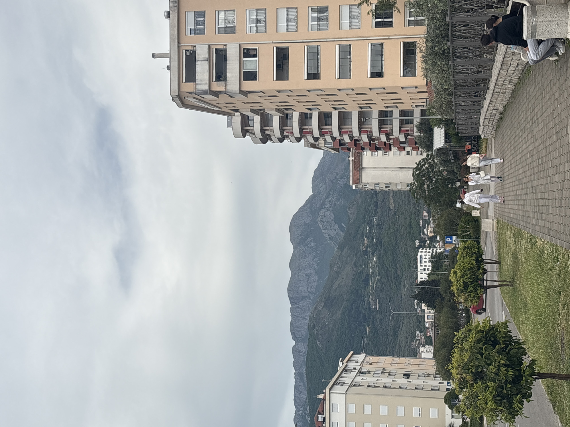 The image shows a street scene in Bar, Montenegro. A tall, light brown apartment building with many windows and balconies is on the left. Mountains rise in the background. A paved sidewalk runs along the right side of the image, lined with trees and grass. Several people are walking or standing on the sidewalk, including a group of three individuals dressed in white and two people sitting on a stone bench near a fence. The sky is overcast. A parking sign can be seen between the trees. A red car is partially visible.