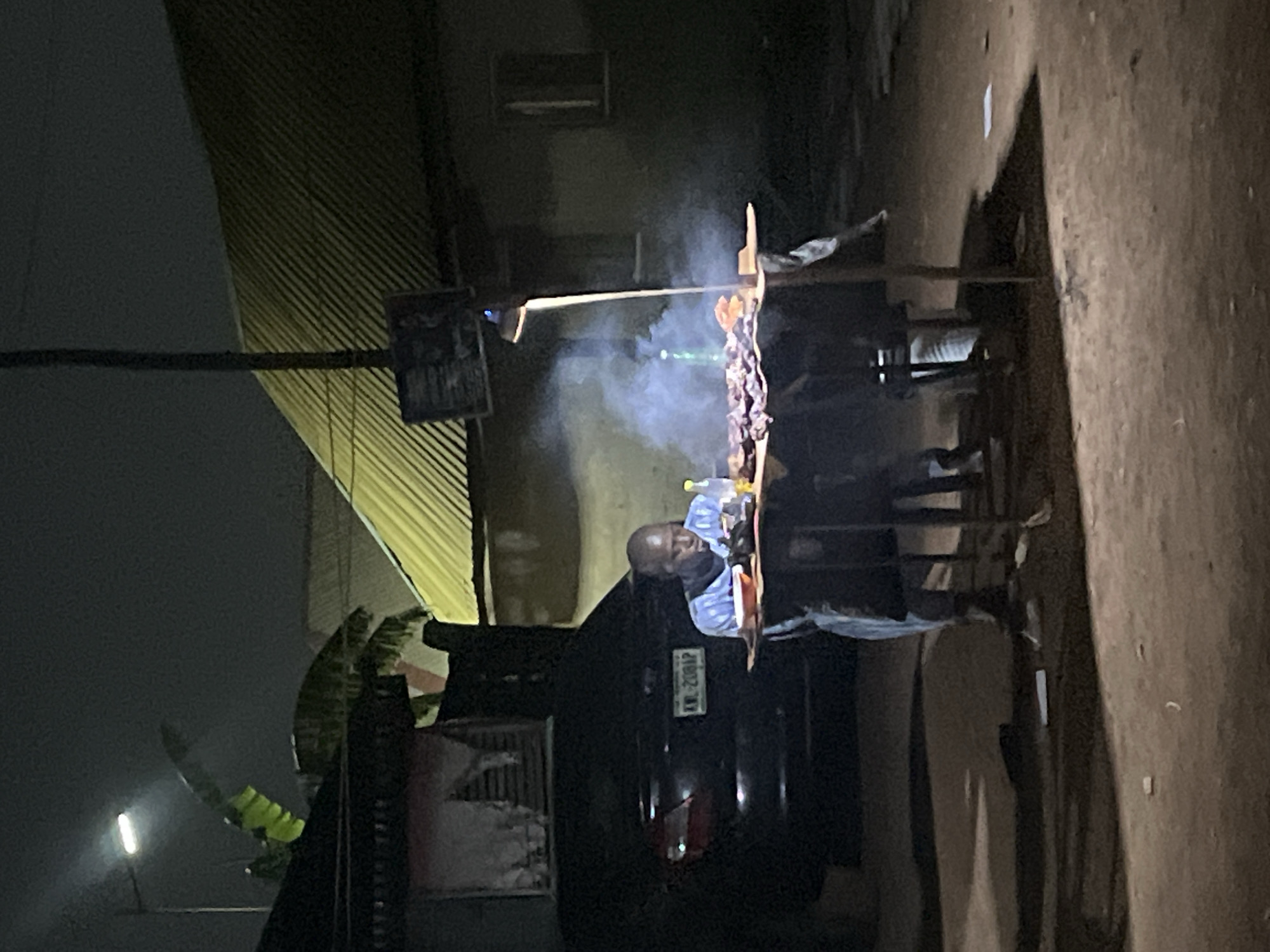At night, in what appears to be a street in Abuja, Nigeria, a man with a bald head wearing a light blue and white striped shirt stands behind a food preparation area. The food is smoking on a grill. The cooking area is adjacent to a black car with a license plate that reads "KW-2381P." A corrugated metal roof overhangs the area. A street light is visible in the distance.