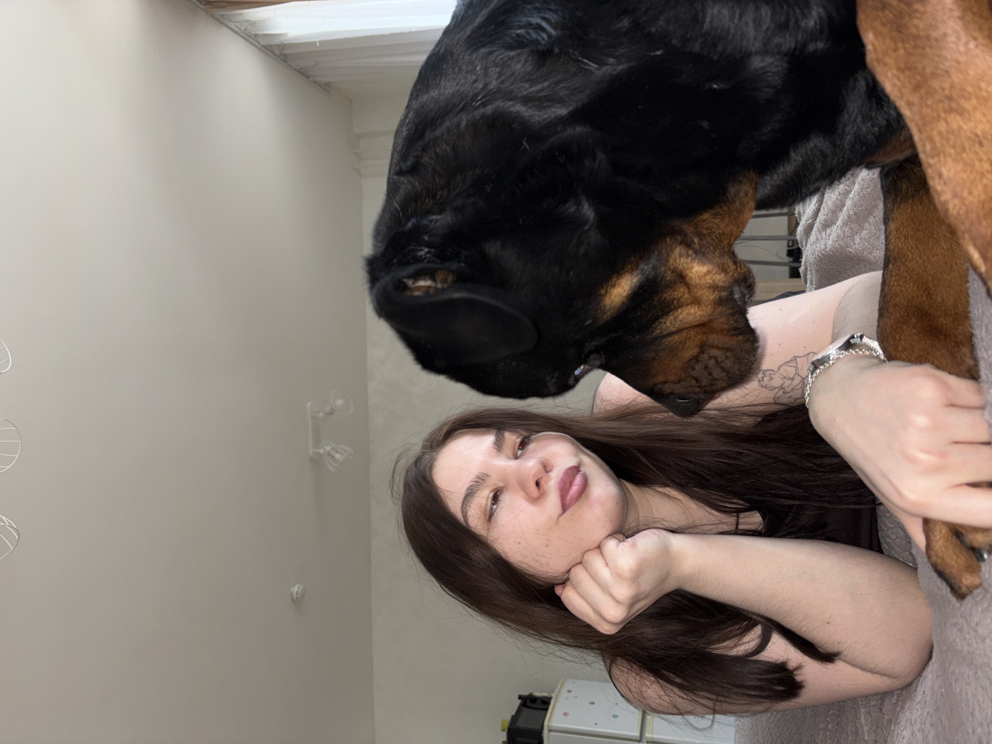 A woman lies on a gray surface, possibly a bed or couch, with a large Rottweiler dog positioned above her, partially obscuring her upper body. The woman has long brown hair and fair skin. Her left hand is placed under her chin, and she looks directly at the camera. She has a silver bracelet on her right wrist. The dog is predominantly black with brown markings around its muzzle and legs. The setting appears to be indoors, with a light-colored wall visible in the background, decorated with abstract white wire decorations and a wall light fixture. A white electrical outlet or appliance is visible in the lower right corner. The location is Perm, Russia.