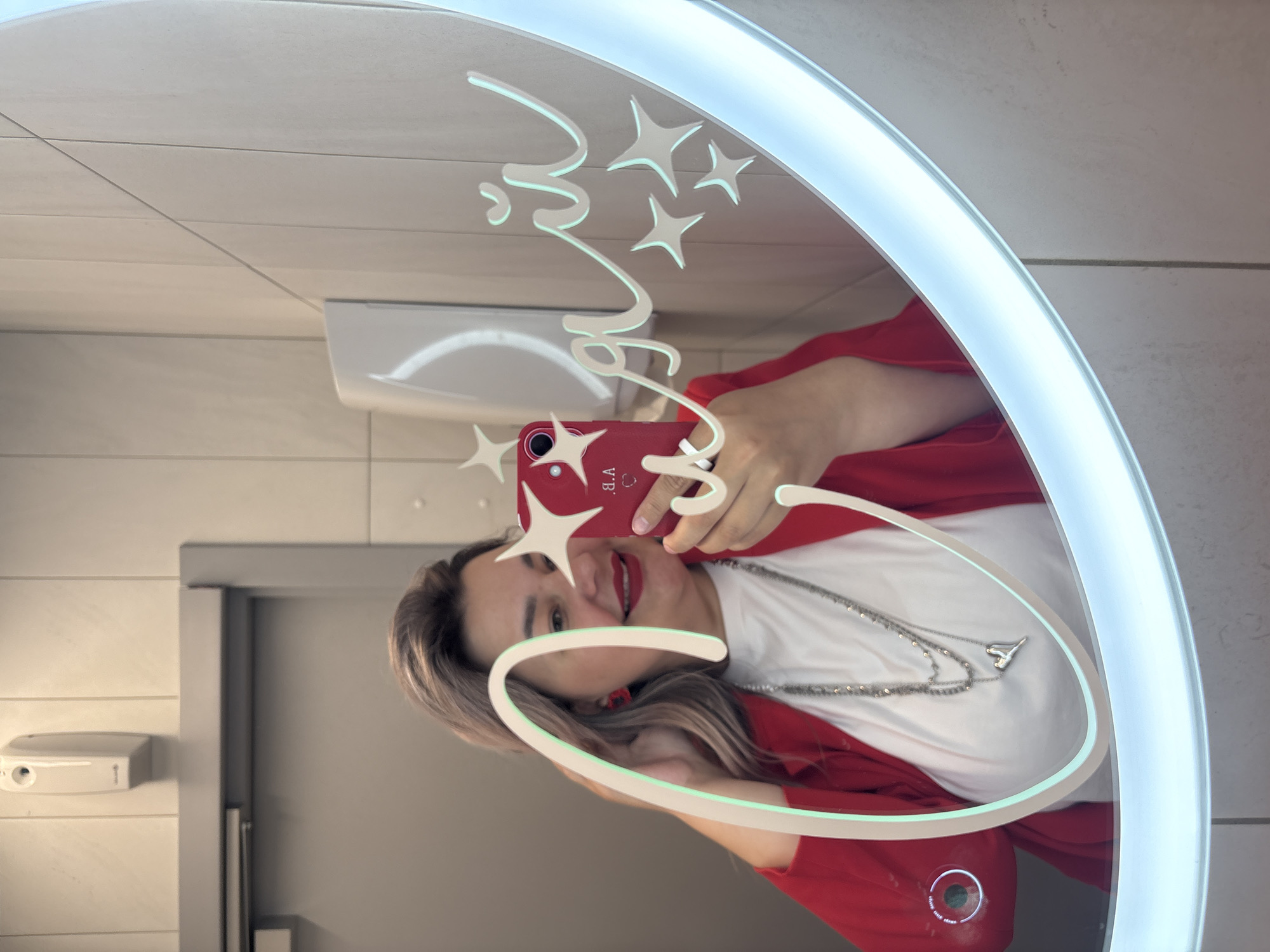 A person is taking a selfie in a brightly lit, oval mirror with designs, possibly in a bathroom or restroom. The person is wearing a red jacket over a white shirt and a multi-strand necklace with a pendant. They are holding a red smartphone up to the mirror, with the phone's camera and flash visible. The mirror has the word "magic" written in a stylized script and several stars etched into the glass above the phone. The background features light-colored wall tiles, a white hand dryer, and a grey door. While it is not possible to pinpoint the location as Birobidzhan, Russia, without further information, the details provided describe the scene and the people and objects within it.