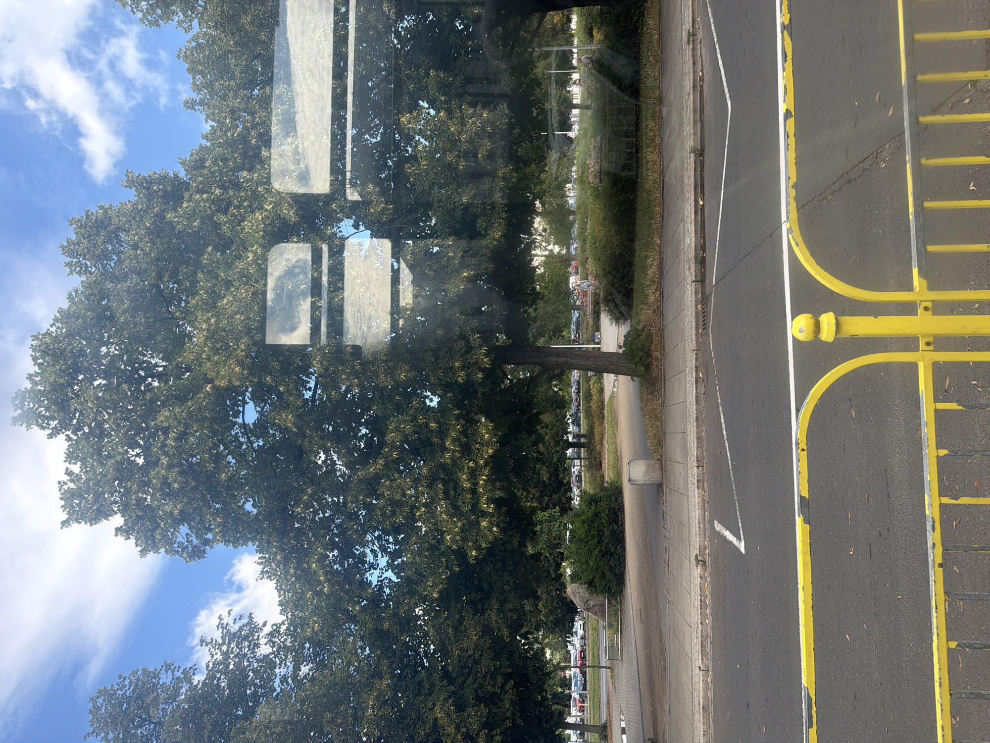 The image shows a street scene with a tree-lined sidewalk and a parking lot in the background. A yellow metal fence is in the foreground, and a road with white lane markings is visible. The sky is blue with scattered clouds. The overall impression is that of a bright, sunny day in an urban environment.
Without additional information or context, it is impossible to determine the specific location in the city of Szczecin, Poland.