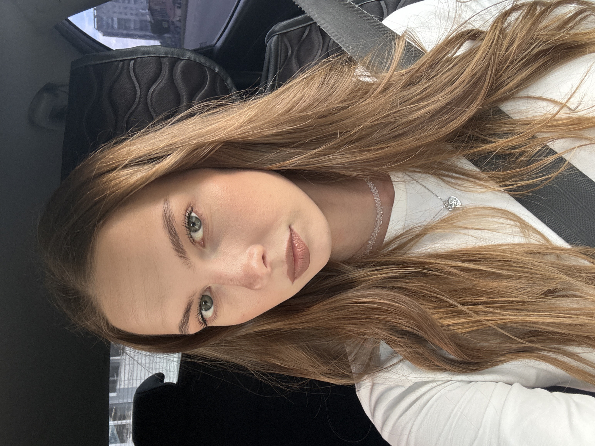 The image shows a young woman with long, reddish-brown hair, wearing a white top and a necklace, inside a car. She is looking directly at the camera with a neutral expression. The background suggests an urban environment, with buildings and a street visible through the car window. The specific location in the city of Perm, Russia, is not identifiable from the provided image.