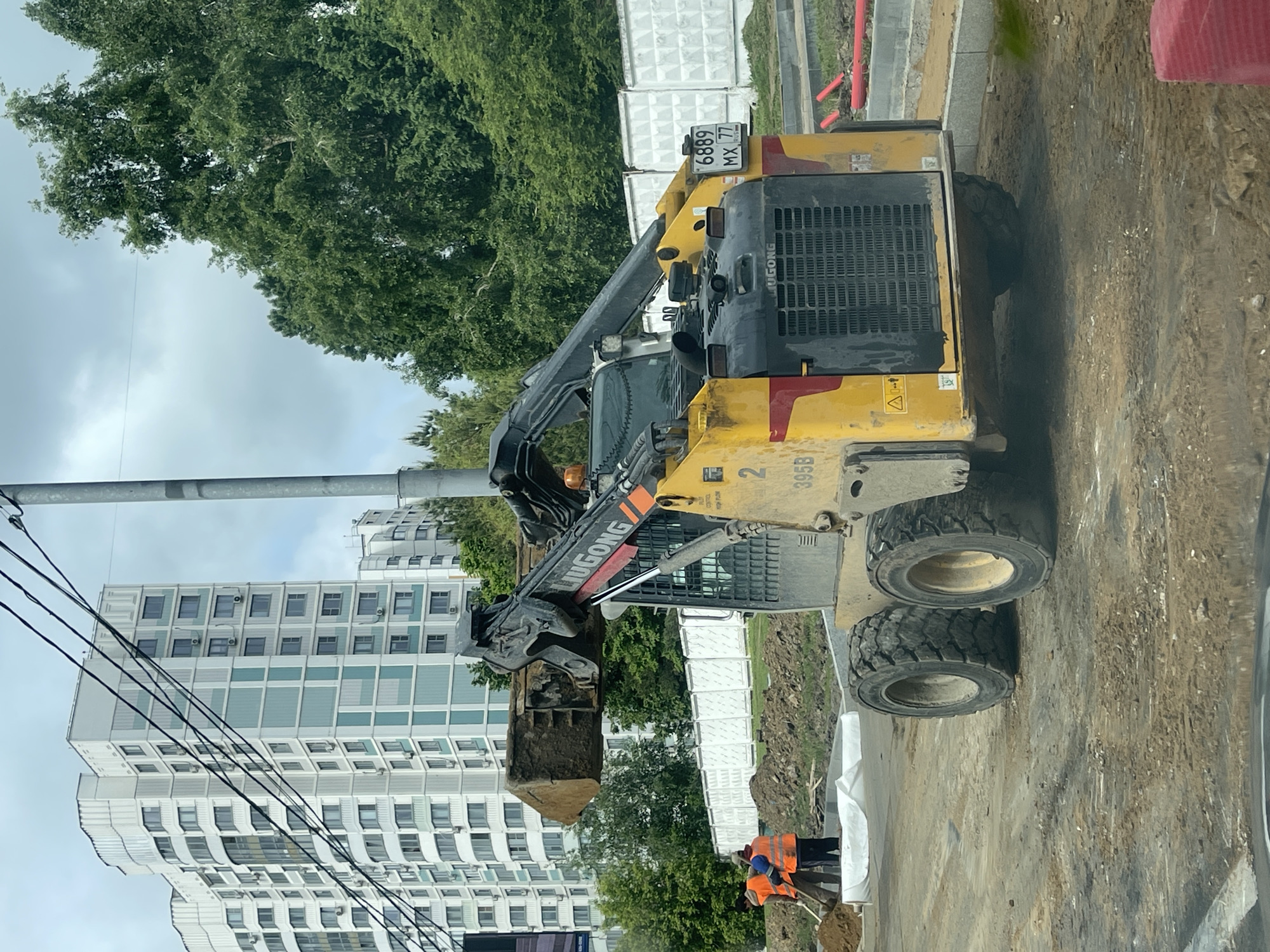 The image shows a construction site in Moscow, Russia, with a Lingong 395B skid-steer loader holding a bucket of dirt. The loader has the license plate 6889 MX 77. Two construction workers in orange vests are visible near the loader. In the background are a tall residential building and trees, with a cloudy sky above. Overhead power lines are also visible.