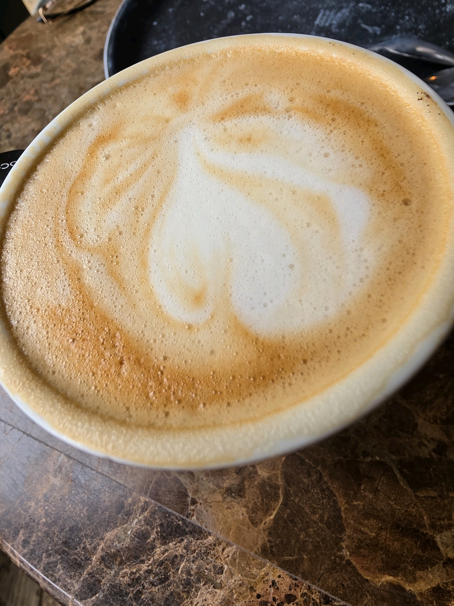 The image shows a close-up, overhead view of a latte in a white ceramic mug. The latte has a heart shape design in the foam, created with darker coffee. The mug sits on a brown, marble-patterned table. In the background, a dark, circular object, possibly a saucer or a placemat, is visible, along with a hint of eyeglasses. The scene is likely indoors, in a cafe or restaurant setting. There are no people or animals clearly visible in the image. The lighting suggests it may be daytime. No specific landmarks or identifying features of West Suffolk are discernible.