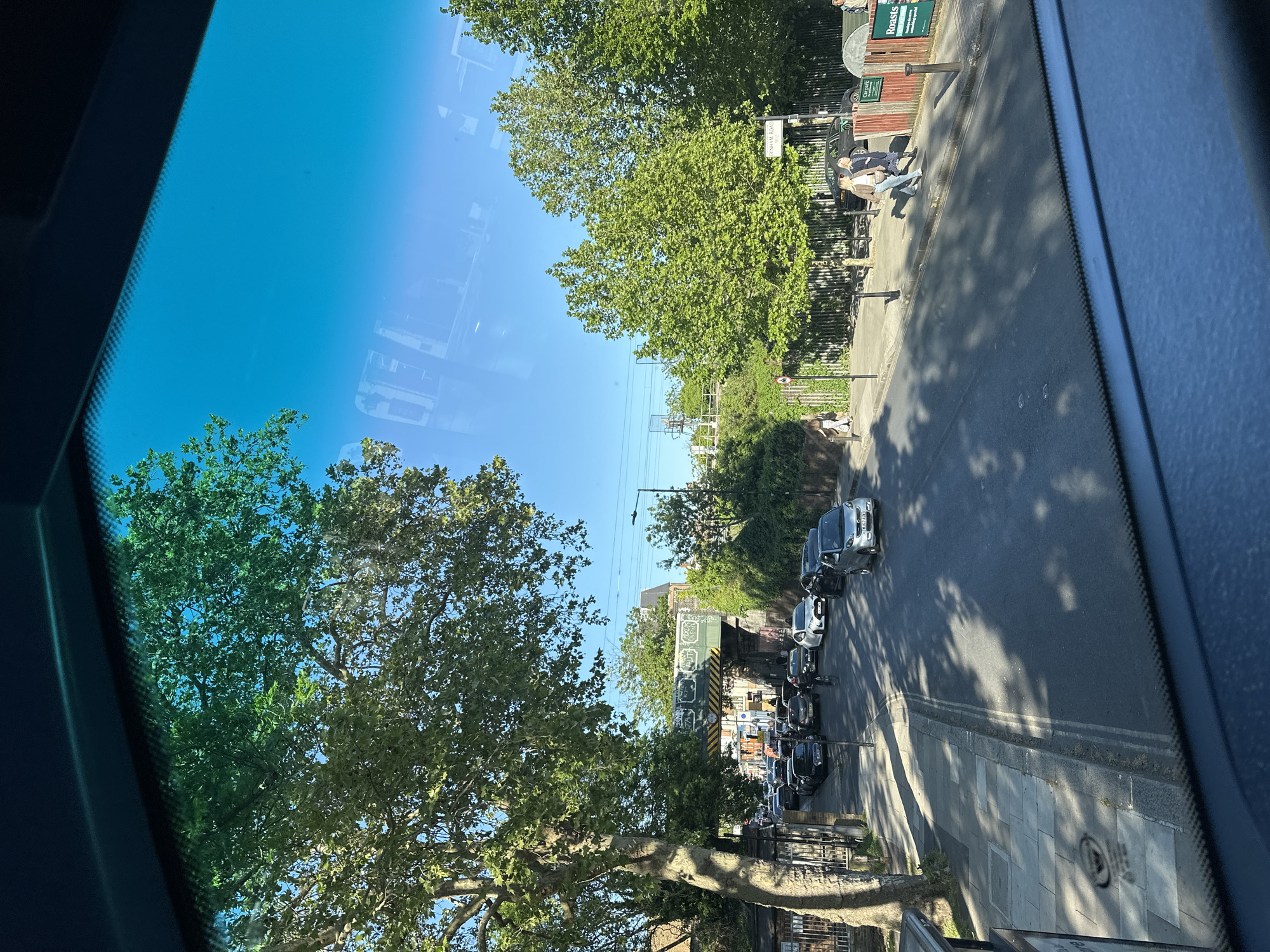 This image captures a bright, sunny urban street scene, likely in London, from an elevated vantage point, possibly inside a vehicle like a bus. The view looks down onto a road lined with parked cars and sidewalks.
The sky is a clear, vibrant blue. Large, mature trees with abundant green foliage dominate both sides of the street, their leaves illuminated by strong sunlight. These trees cast prominent, dappled shadows across the pavement and road, suggesting it is either morning or late afternoon.
On the right side of the street, numerous cars are parked in a row, including a silver hatchback, a dark SUV, and various other sedans. Along the left side, a paved sidewalk features several pedestrians. In the middle ground, two individuals are standing near a wooden fence, one dressed in a light-colored jacket and jeans, and the other in darker clothing. Further down the street, more people can be seen walking or standing.
Notable objects include a street sign clearly bearing the text "INHAM ROAD," a partially visible sign indicating "Car Park," and another sign mentioning "Roasts," suggesting a food establishment. A red circular road sign with a white diagonal line (typically signifying "No Waiting") is also visible. In the background, a railway bridge or underpass, painted with green and yellow stripes and adorned with some unreadable graffiti, spans the street. The overall setting depicts a typical, lively city street on a clear day.