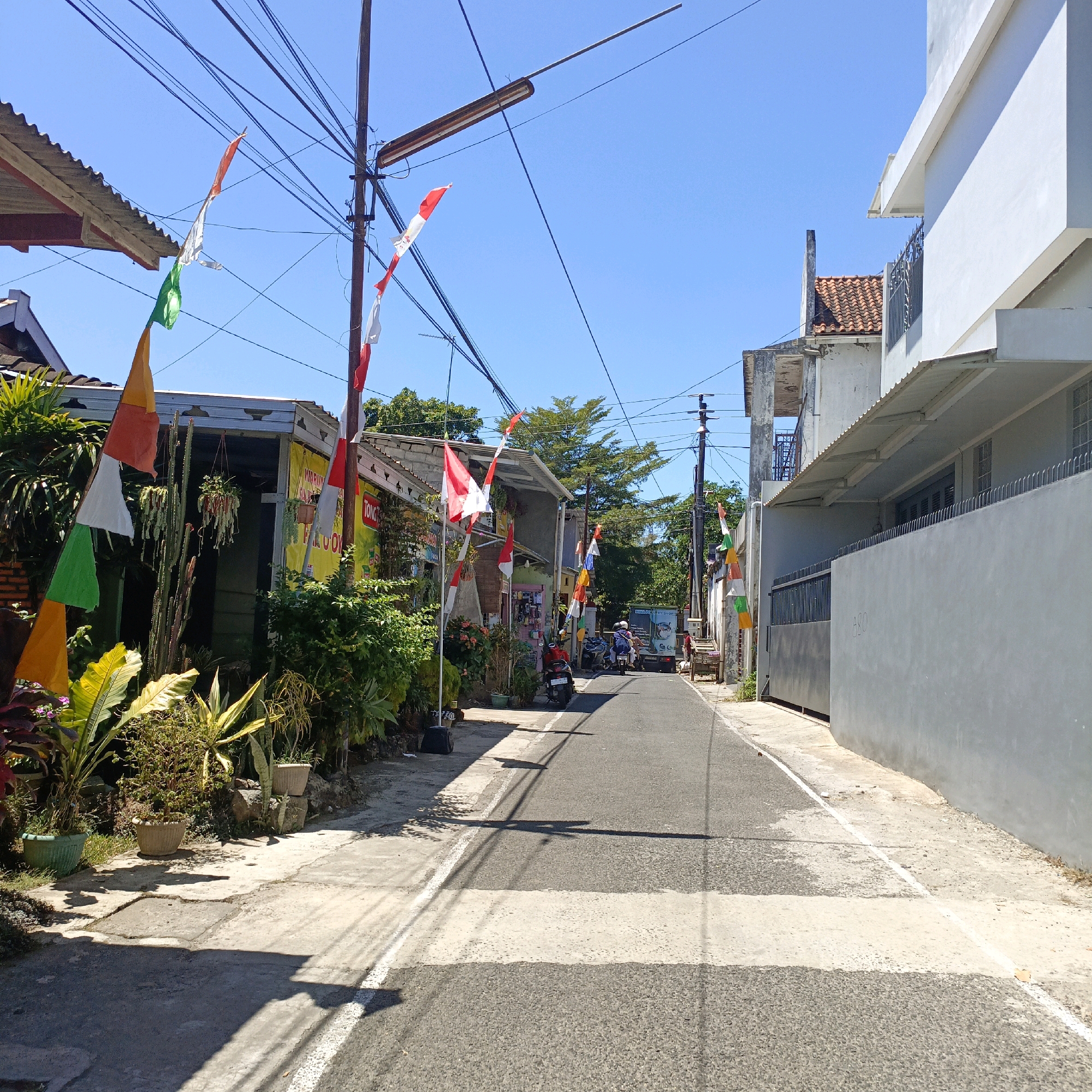 A narrow, paved road in Salatiga, Indonesia, is lined with buildings and lush vegetation on both sides, under a clear blue sky. Numerous national flags of Indonesia, red and white, are strung along the street, indicating a national celebration or commemoration. On the left, a building with a corrugated metal roof features potted plants and dense greenery. On the right, a modern, white building with a grey wall and a metal fence dominates the view. Several motorcycles and a small truck are parked or moving along the road. The scene is bathed in bright sunlight, casting shadows on the pavement.