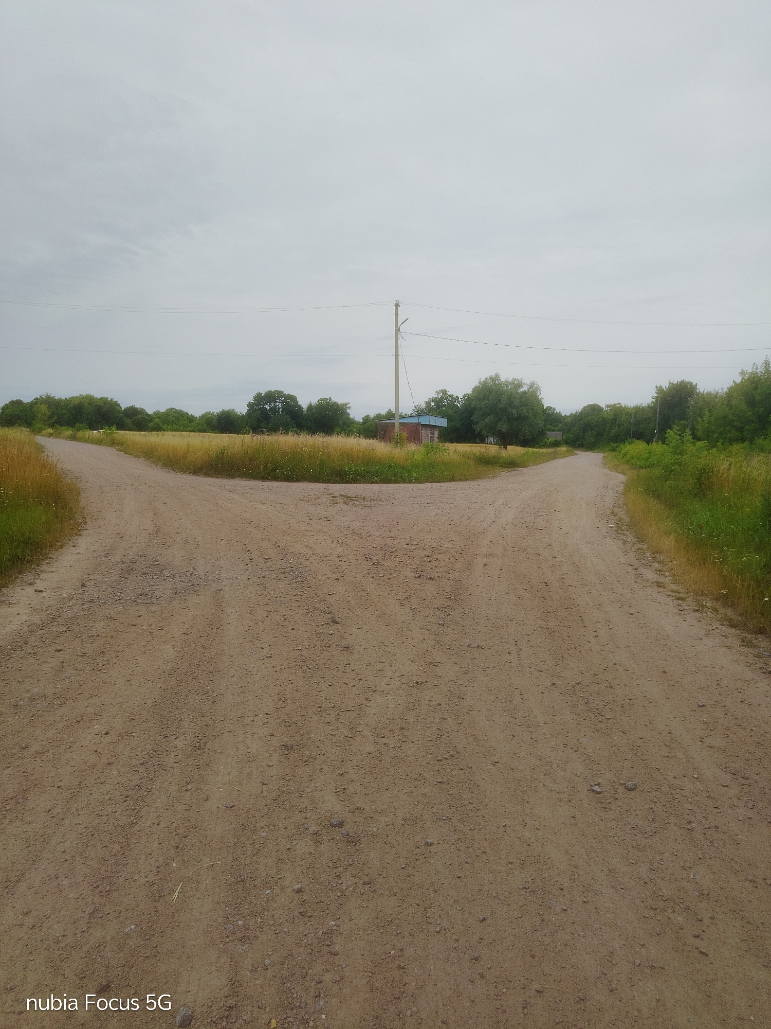 An overcast day presents a fork in a dirt road. The road is a light brown color with scattered small stones and pebbles. Tall, dry grass lines the edges of the road on both sides. In the distance, a line of green trees forms a backdrop across the horizon. To the left of the road, a low-lying field of dry, golden grass is visible. To the right, a darker green patch of grass and foliage is apparent. In the center background, a small, brick building with a blue roof sits near a wooden utility pole with wires extending horizontally. The sky is a uniform, light gray, suggesting cloudy weather. The lower left corner of the image displays the watermark "nubia Focus 5G".