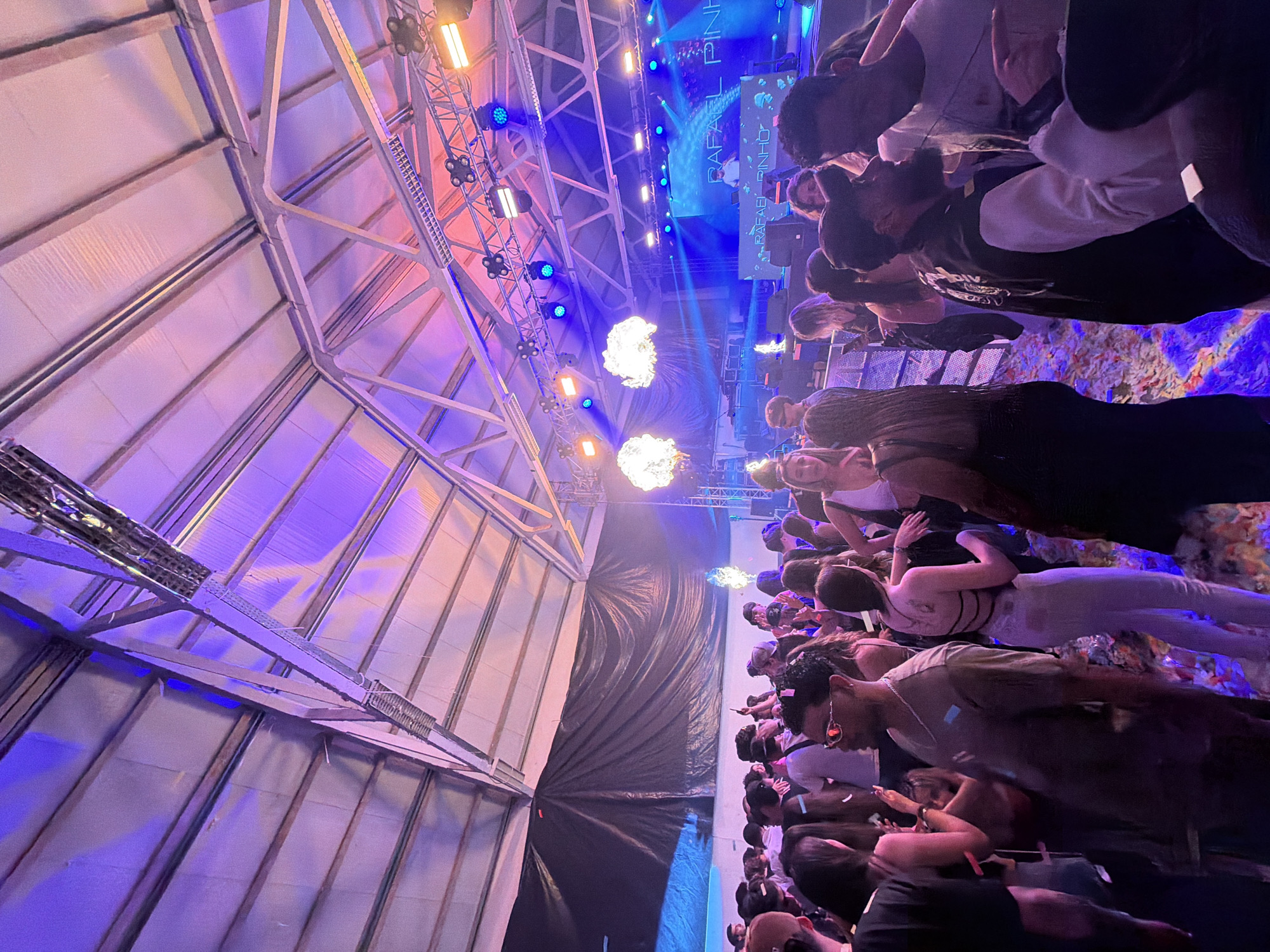 The image shows an event at a concert venue, likely in Bragança, Portugal, judging by the text displayed on the stage's screen. A crowd of people is looking towards a stage with bright lighting and flame effects. The structural framework of the indoor venue is exposed, revealing the ceiling and support beams. The color scheme includes blue, purple, and orange hues from the stage lighting. People are standing close together, and some may be recording the event.