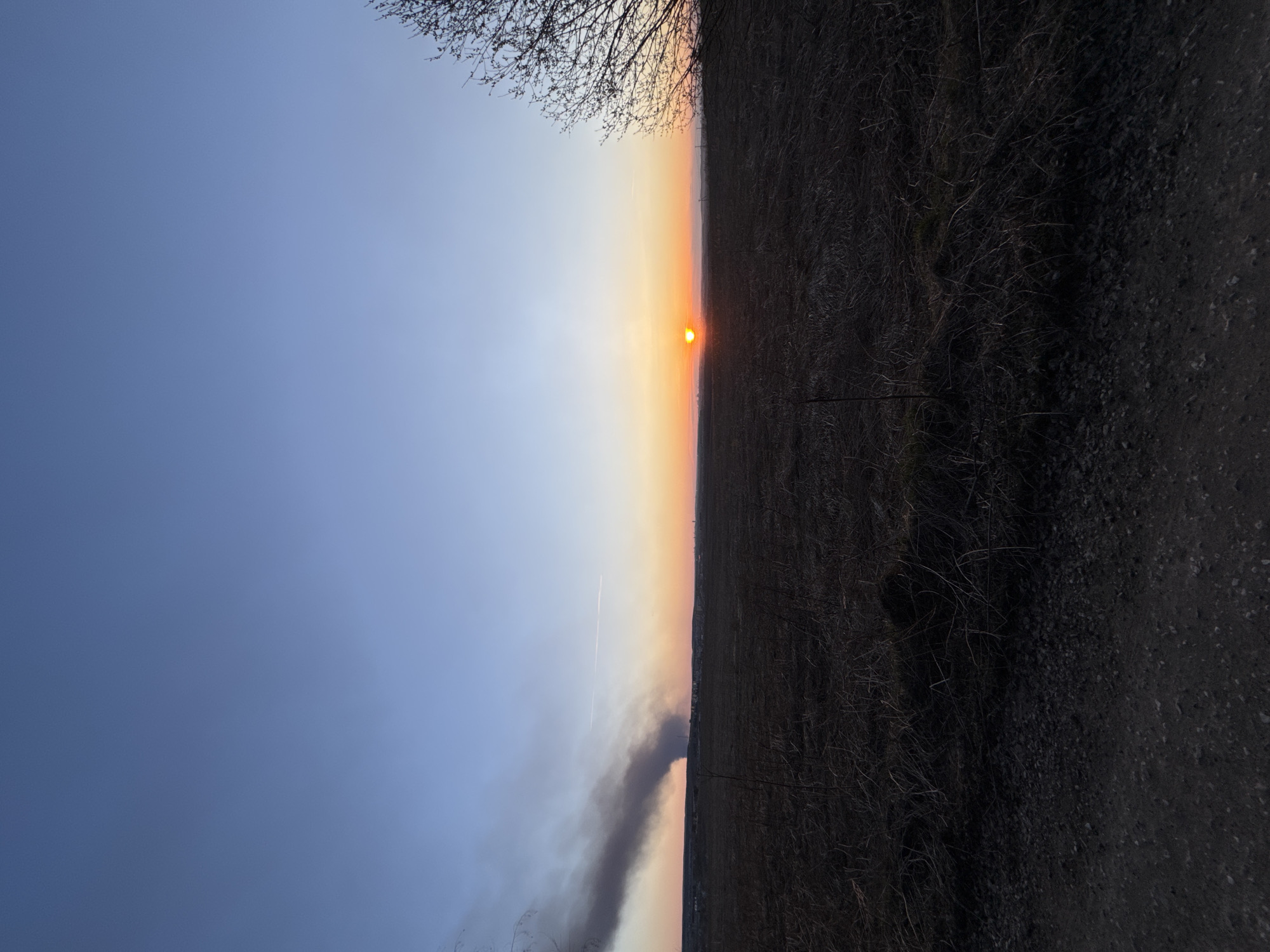 The image depicts a landscape at sunset, likely near Kochkino, Russia, although specific urban features are not discernible. The horizon is bisected by a band of orange and yellow light indicating the setting sun. The sky transitions from light blue to orange, with a contrail of a jet visible. On the ground to the right is a dark field of dormant vegetation leading to a path or road. To the left, a plume of dark smoke rises into the sky. Some bare branches of a tree are visible in the upper right corner. There are no people or recognizable man-made objects visible apart from the smoke and possible contrail. The environment appears rural and possibly dry.