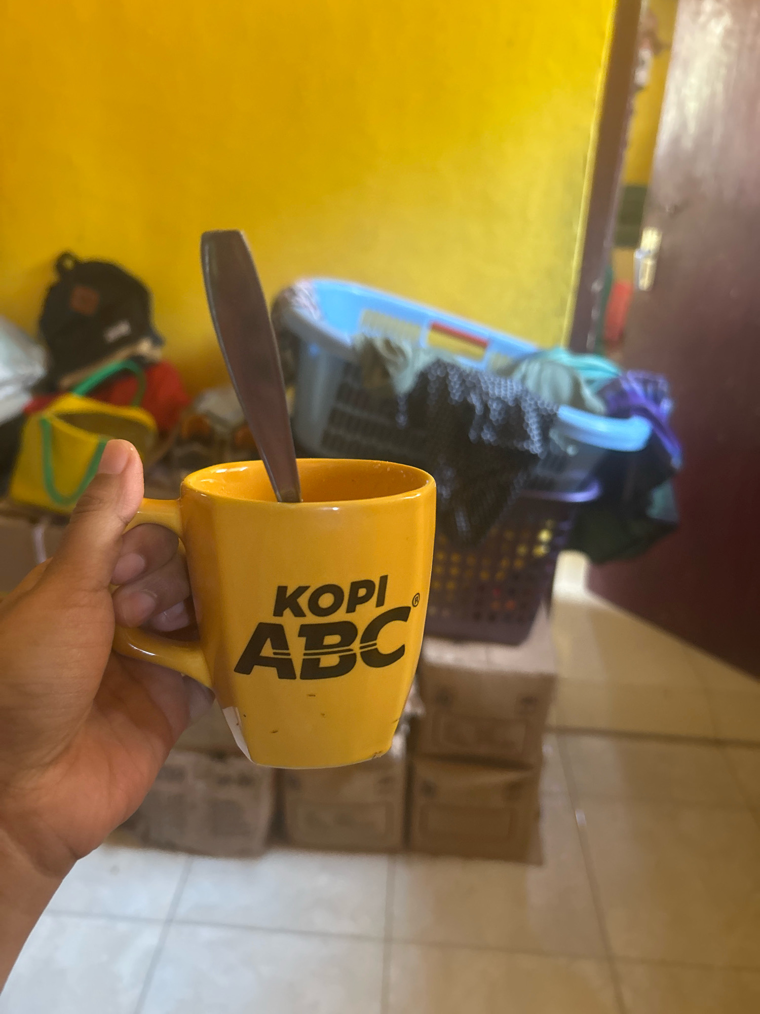A person's hand is holding a yellow mug with "KOPI ABC" written on it in black lettering. A metal spoon is inside the mug. The background is a yellow wall with some laundry in a blue and purple basket. There are also some boxes and a yellow bucket visible. The scene appears to be indoors, possibly in a home in Cihaurbeuti, Indonesia.