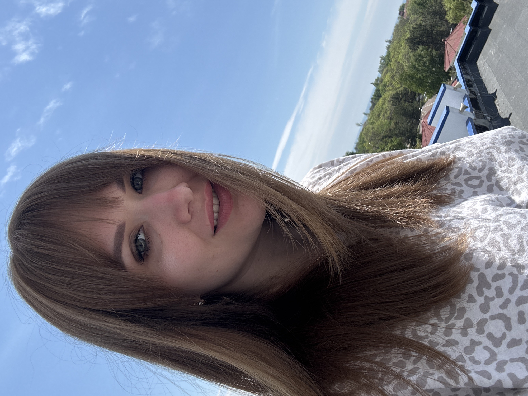 A young woman with brown hair and bangs is smiling into the camera. The photo appears to be taken outdoors in Yevpatoriya, Ukraine, based on the architecture visible in the background which includes white buildings with blue trim and red tile roofs. The sky is blue with some clouds. The woman is wearing a light-colored shirt with a print.