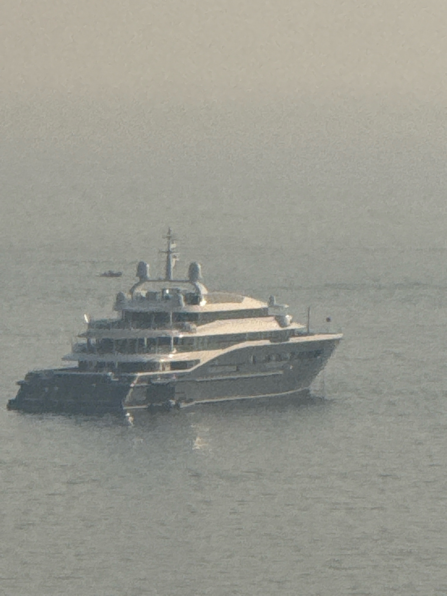 A large, dark grey superyacht is anchored in calm, grey water, likely in the Bay of Kotor near Boreti, Montenegro. The yacht has multiple decks, a prominent mast structure at the rear, and a sleek, modern design. In the distance, a smaller, dark boat is also visible. The sky is overcast, creating a hazy, diffused light that gives the scene a muted appearance.