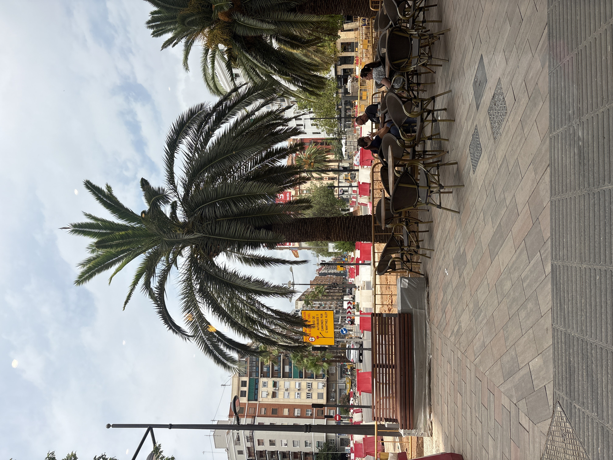 The image depicts a street scene in Valencia, Spain, likely in an urban area undergoing construction. A prominent palm tree dominates the foreground, with its fronds partially obscuring the view of the sky, which is overcast with scattered clouds. To the left, a street lamp stands next to a wooden bench covered with plastic sheeting. Across the street, a multi-story building with an orange-brick facade is visible, along with other buildings of varying architectural styles. The street itself is blocked off with red and white construction barriers, and there are signs indicating detours. In the mid-ground, there is a cafe with tables and chairs set up outdoors. Three people are seated at one of the tables, engaged in conversation. The ground is paved with rectangular bricks.