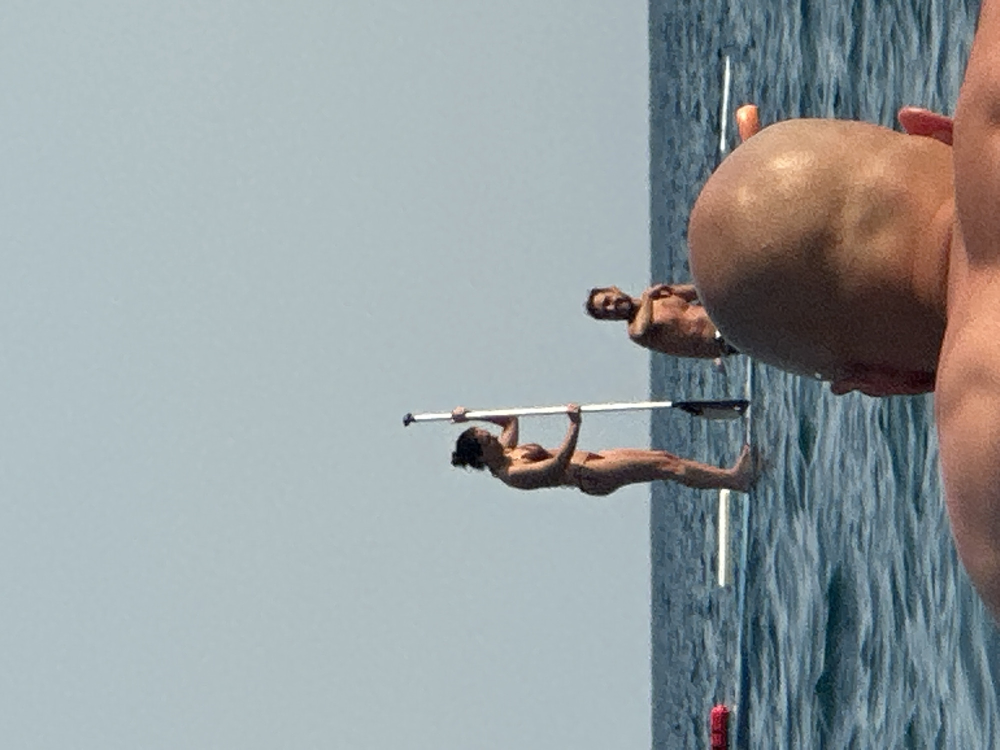 The image depicts a bright, sunny day at sea, likely near Boreti, Montenegro, given the coastal landscape and calm waters. In the foreground, the back of a bald person's head and shoulders are visible, facing away from the camera, suggesting they are also on a watercraft or in the water. In the middle ground, two individuals are engaged in paddleboarding. A woman, standing on a paddleboard, holds a paddle vertically. She is wearing a bikini. Beside her, a man is seated on another paddleboard, looking towards the woman. The sea is calm with small waves, and the sky is a clear, pale blue. Several buoys are visible in the water, one red and one orange, along with a white object that might be another paddleboard or a flotation device. The overall scene suggests a leisurely outdoor activity in a coastal environment.
