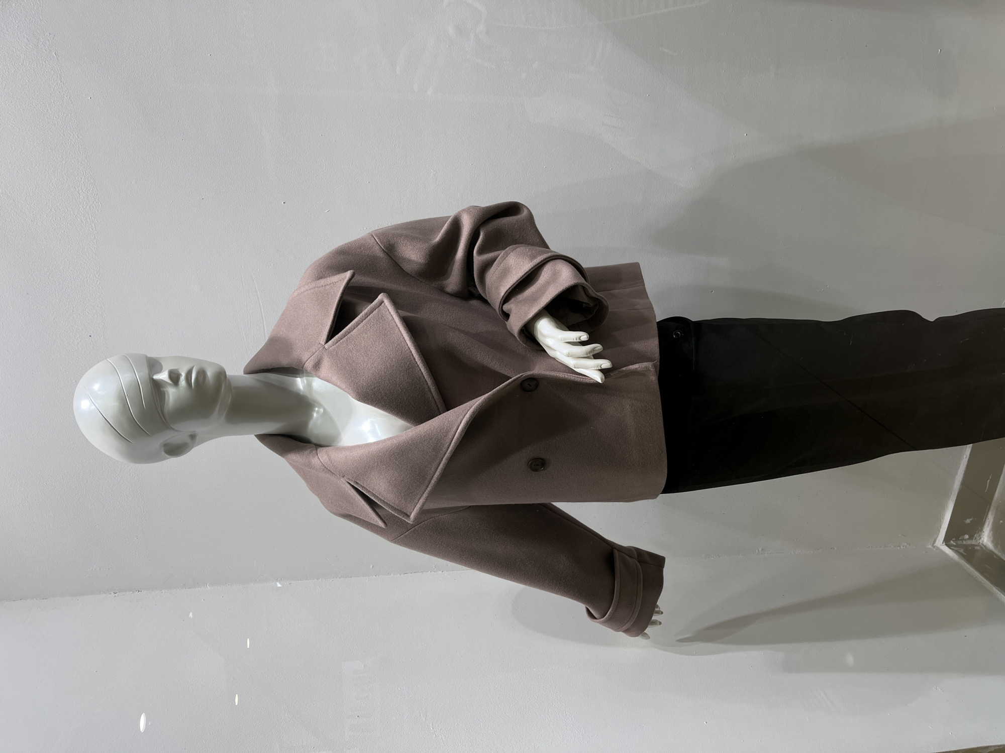 A mannequin displays clothing in a retail setting. The mannequin, composed of a shiny white material, has a faceless, stylized head and is angled to the left. It wears a light brown, double-breasted jacket with broad lapels. The jacket's sleeves are slightly crumpled, suggesting movement or wear. Below the jacket, the mannequin is dressed in dark-colored trousers. The setting appears to be a storefront window, with a bright white wall in the background that reflects light. The floor is also white and reflective, indicating a clean and modern display area. The location is in the city of Unknown, Unknown.