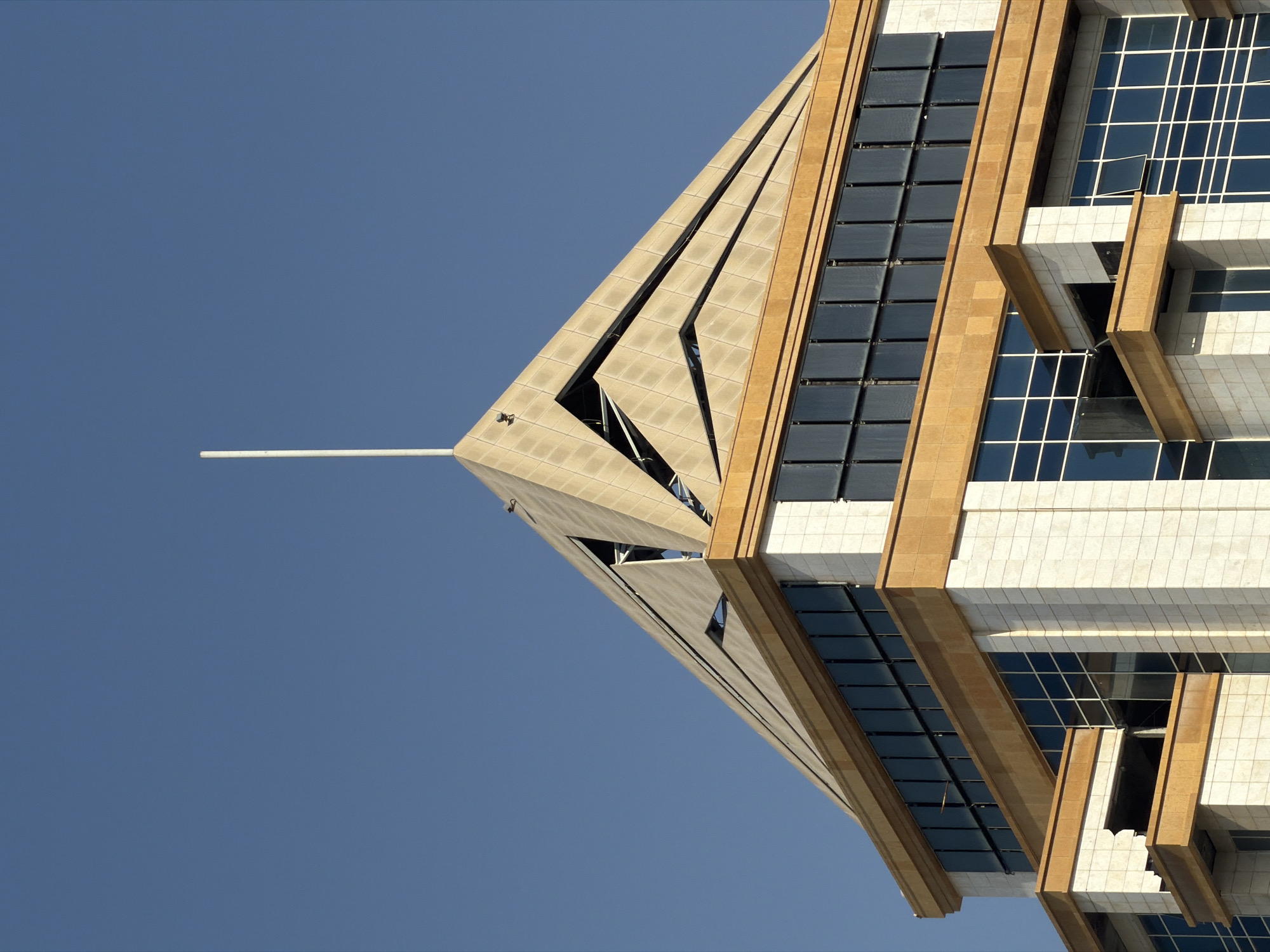 A close-up, slightly upward shot shows the top of a modern skyscraper against a clear blue sky. The building has a unique, angular design with a pyramidal top. The upper section of the pyramid is clad in light beige tiles arranged in a diamond pattern, with deep triangular recesses revealing internal structures. A thin, white rod or antenna extends vertically from the apex of the pyramid. Below the pyramidal section is a level with a band of dark, rectangular panels, possibly solar panels, set against a prominent band of tan-colored stone. The lower visible levels of the building are clad in light-colored stone blocks and feature large, dark-tinted windows framed by white mullions. Some of the windows are open. The architectural style suggests a contemporary, possibly corporate or governmental building. The scene is static and appears to be an architectural photograph or a detail shot of the building's facade.