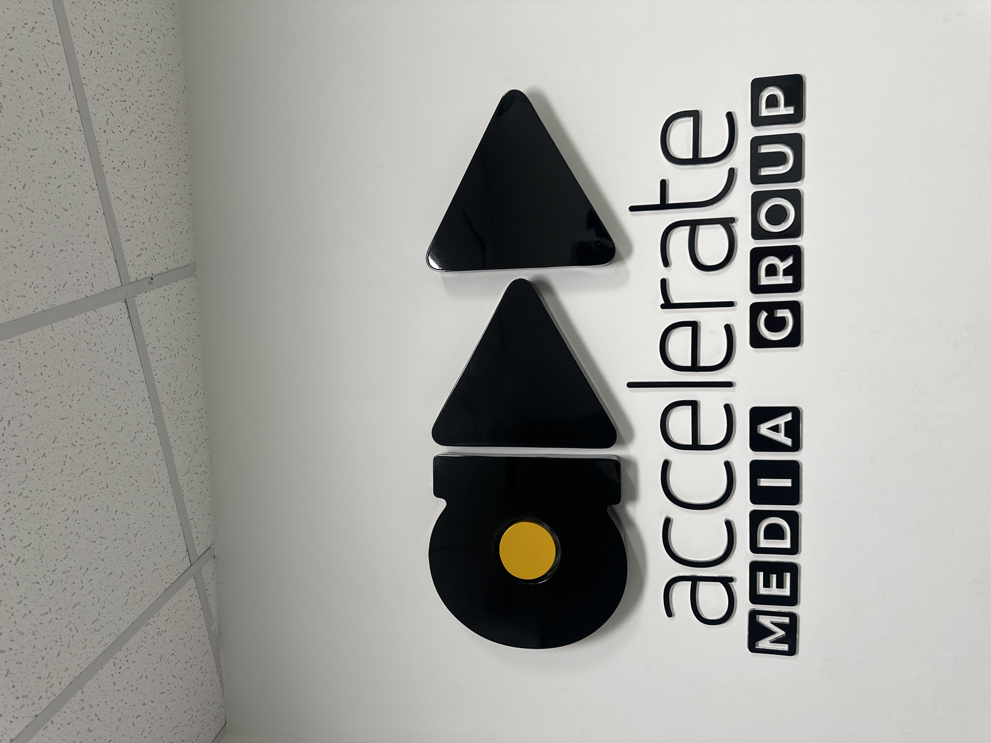 The image shows a wall sign displaying the logo and name "accelerate MEDIA GROUP". The logo features two black triangle shapes stacked vertically above a black circle with a yellow center. The name is written in black letters to the right of the logo. The sign is mounted on a white wall. The left side of the image shows a suspended ceiling with a grid pattern. The location of the image is not evident.