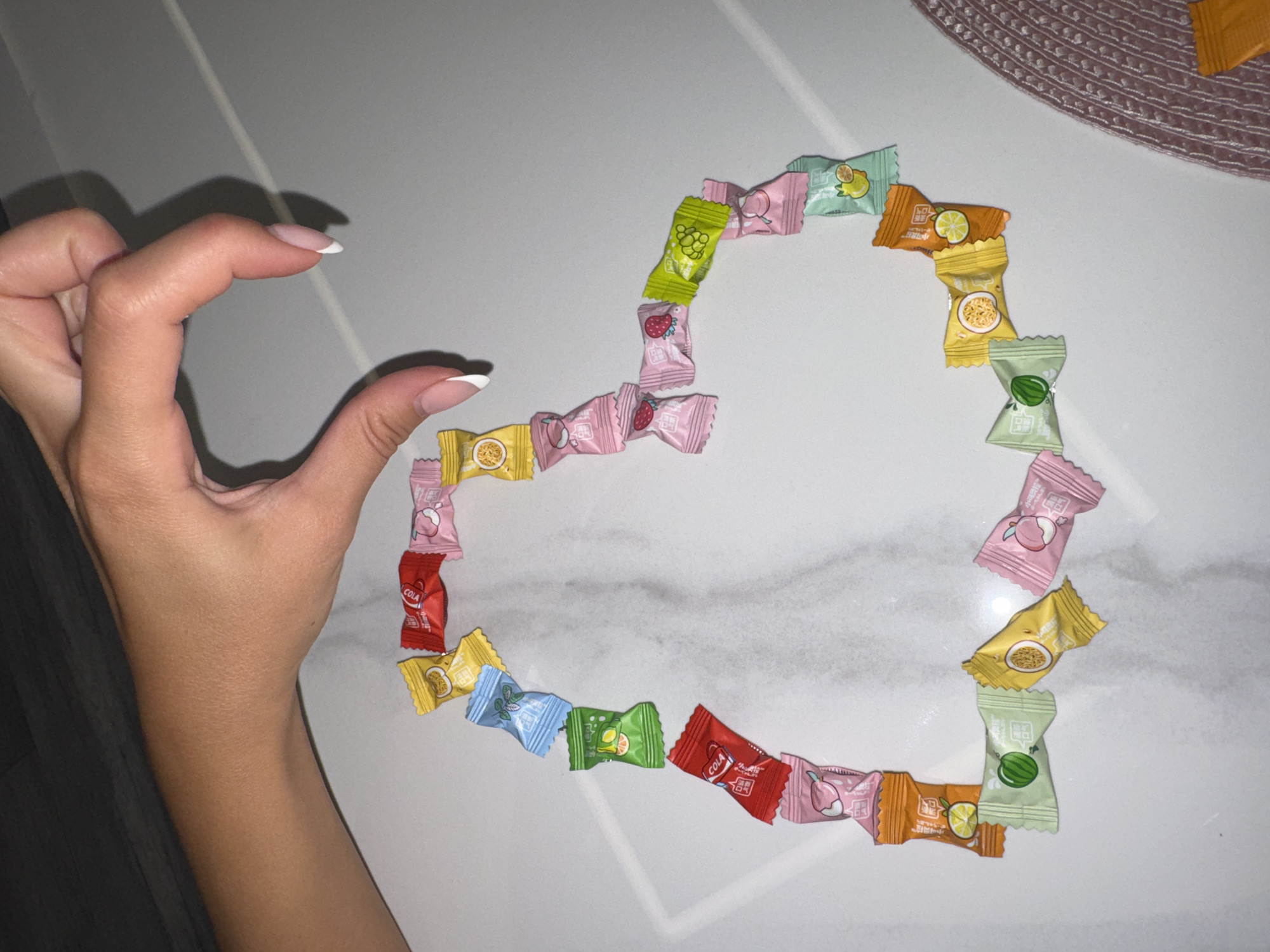 A left hand with manicured nails is positioned near the upper left of the frame, appearing to form the left curve of a heart shape. A heart shape is outlined by small, individually wrapped candies of various colors and fruit designs, situated on a white, possibly marble or granite, surface. The surface appears to be a table. A small portion of a circular woven mat is visible in the upper right corner.
There is no evidence to locate the image at Burgas, Bulgaria.