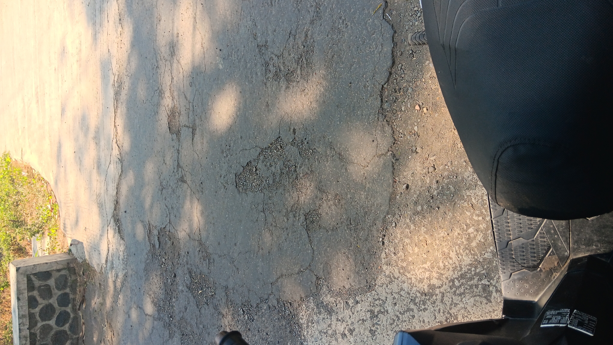The image is a close-up, high-angle shot of a damaged asphalt road with shadows cast by unseen trees. In the foreground, the black seat and part of the body of a scooter are visible, suggesting the photo was taken from the perspective of someone riding or sitting on the scooter. The road surface is cracked and shows several potholes, with loose gravel and debris scattered around. To the left, a concrete structure with dark stones is partially visible, bordering the road. Beyond this, overgrown vegetation hints at a rural or semi-urban environment. The lighting suggests it is daytime, with the sun creating distinct patterns of light and shadow on the road. The location is identified as Gemenggeng, Indonesia.