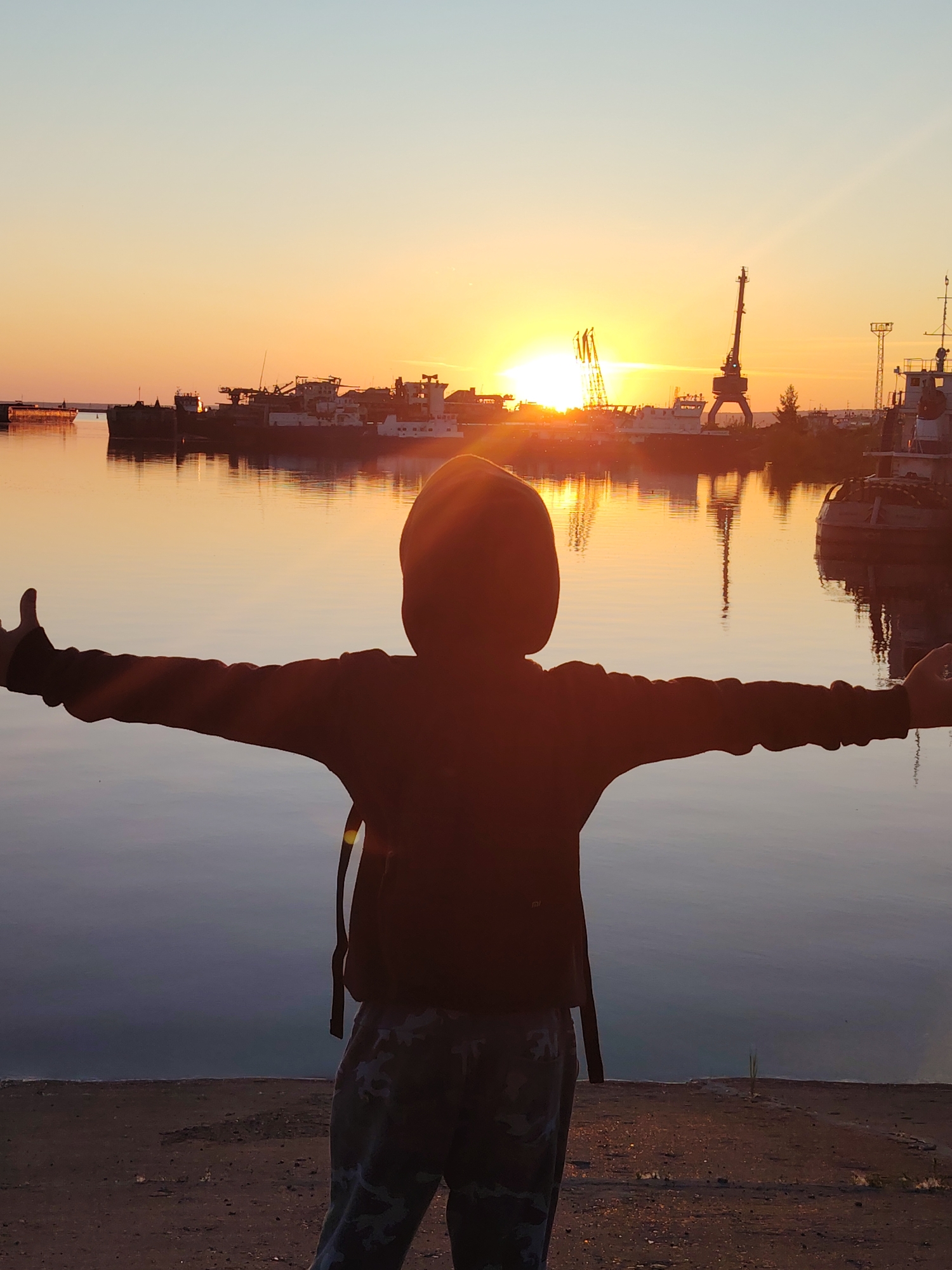 A person in a dark hoodie and camouflage pants stands with arms outstretched towards a sunset over a body of water, possibly a harbor or river in Chaikovsky, Russia. The sun is low on the horizon, casting a warm orange glow across the sky and reflecting on the water. In the background, several ships and cranes are visible, silhouetted against the bright sky. The person appears to be facing away from the camera, with their back to the viewer. The foreground shows a rough, sandy or gravelly surface.