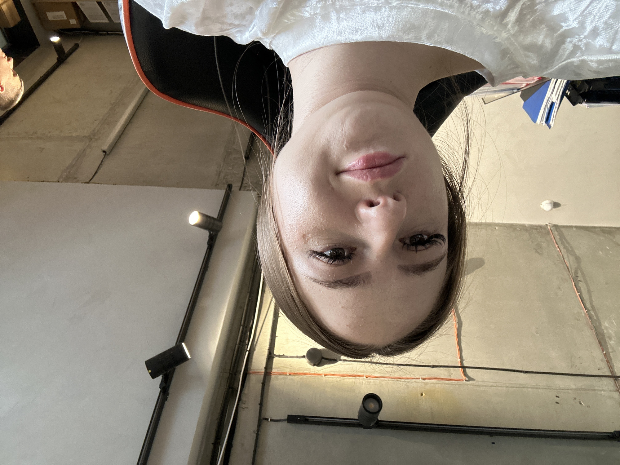 A close-up, selfie-style medium shot captures a young woman from the upper chest to her forehead. She is looking directly at the camera with a neutral expression. The woman has light skin, dark eyebrows, and long, dark eyelashes. Her lips are slightly parted and appear to have a subtle pink tint. She is wearing a white, textured top. Her hair is light brown and appears to be pulled back. The background is an interior space with a concrete ceiling that has exposed conduits and track lighting. There are also some files and office supplies visible in the lower left quadrant of the image. A portion of a black and orange gaming chair is visible on the right side of the frame.