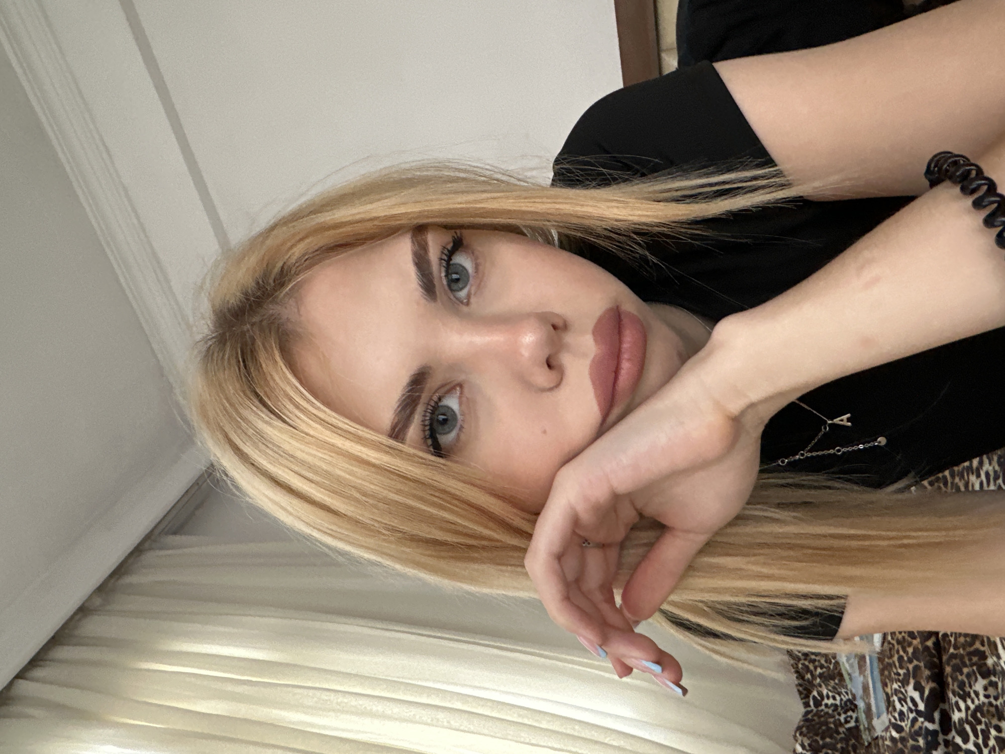 A young woman with blonde hair is resting her head on her hand. She has fair skin and blue eyes. She is wearing a black top and a silver necklace. Her nails are painted with a light blue color. A black hair tie is on her wrist. The background includes a white wall and a beige curtain. Part of a leopard print fabric is also visible.