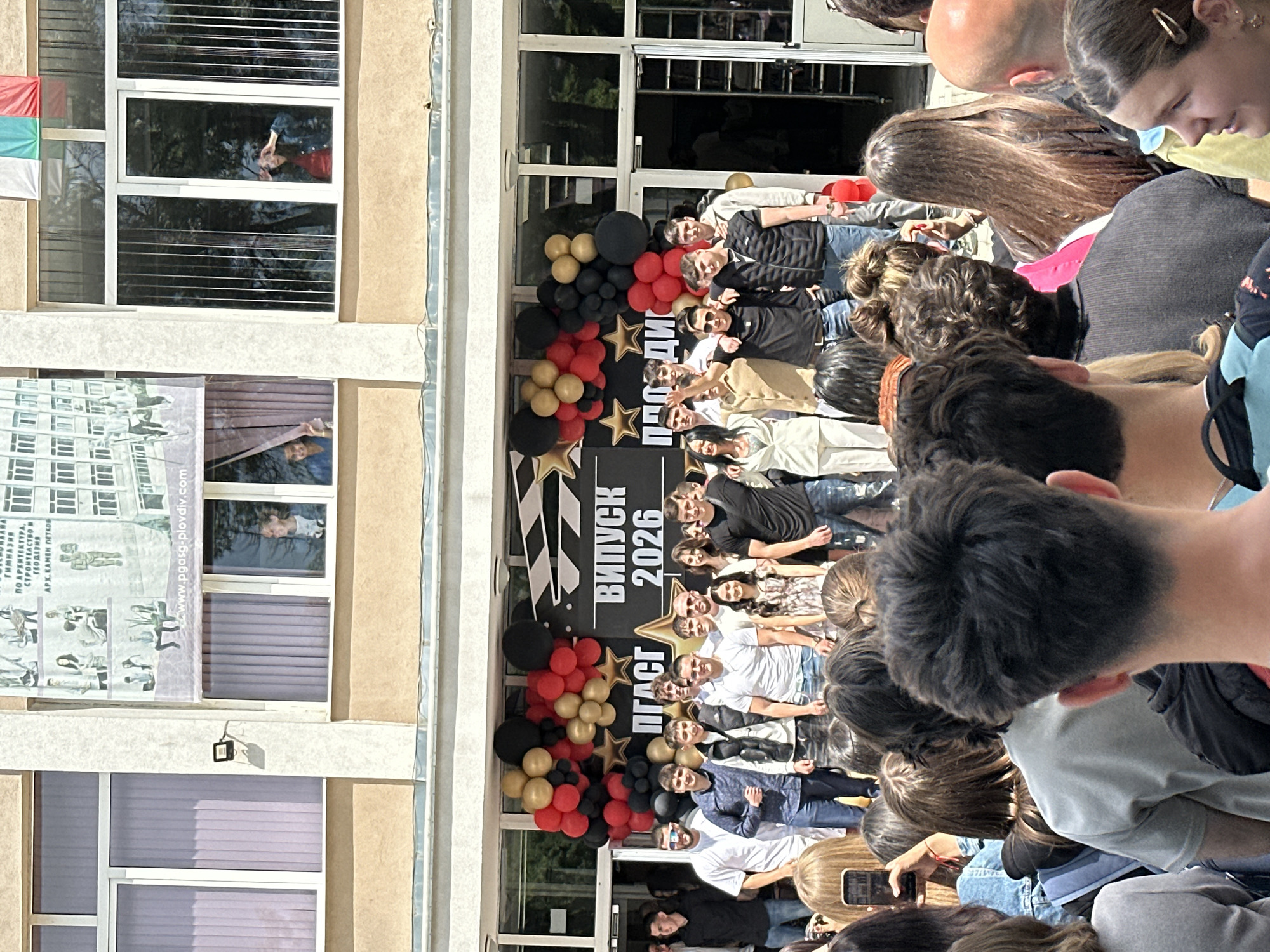 The image depicts a graduation ceremony taking place in Plovdiv, Bulgaria. Graduates are gathered in front of a building adorned with balloons, a banner reading "ВИПУСК 2026" (Graduation 2026), and gold star decorations. The building features windows, one of which shows a person looking out. A banner is hung on the side of the building displaying pictures, possibly related to the school. Observers are present, some recording the event with their phones. A Bulgarian flag is visible hanging in the upper left.